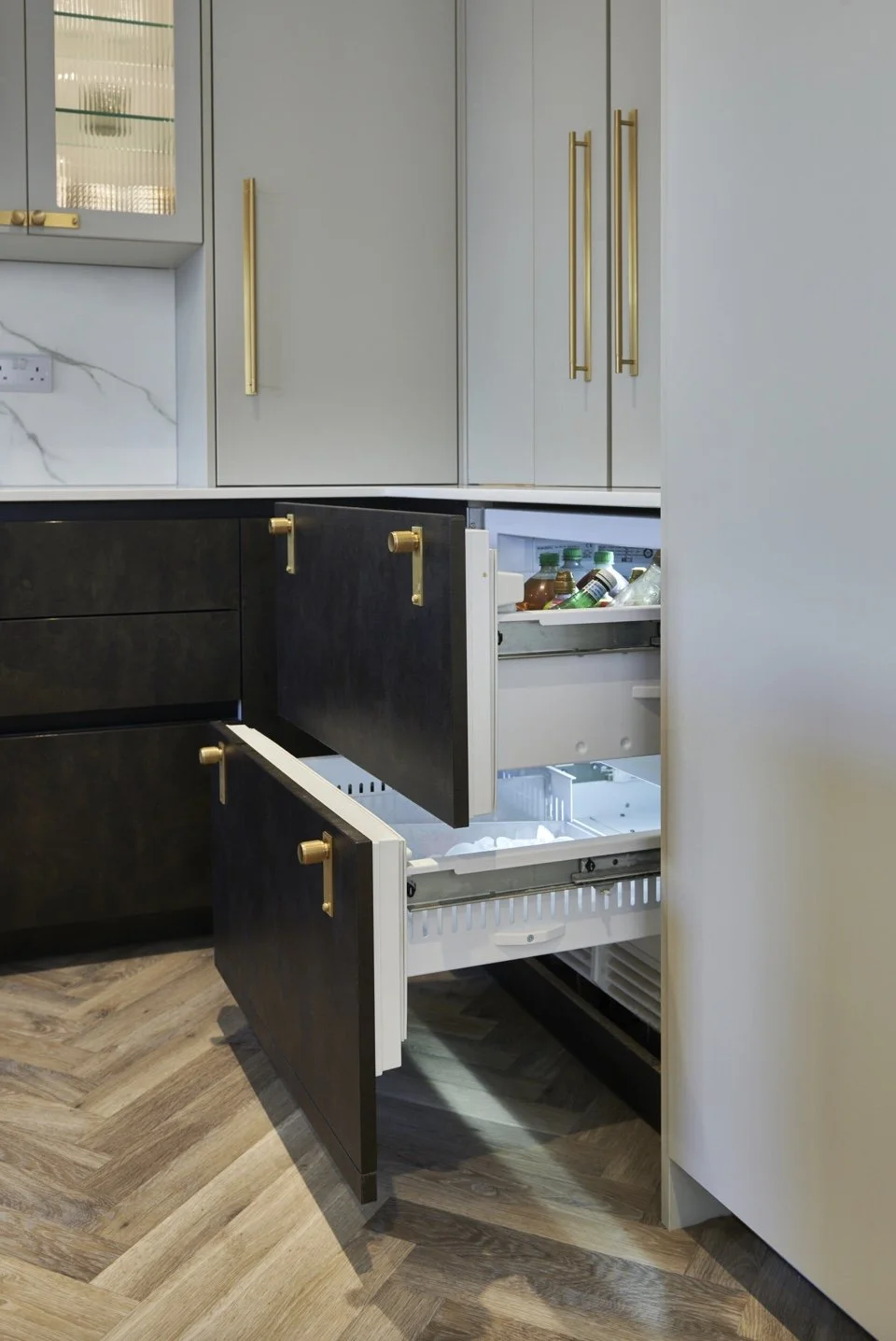 Open kitchen cabinets with a mini fridge containing bottled drinks, modern gray and black cabinetry with gold handles, marble backsplash, and wood-patterned floor.