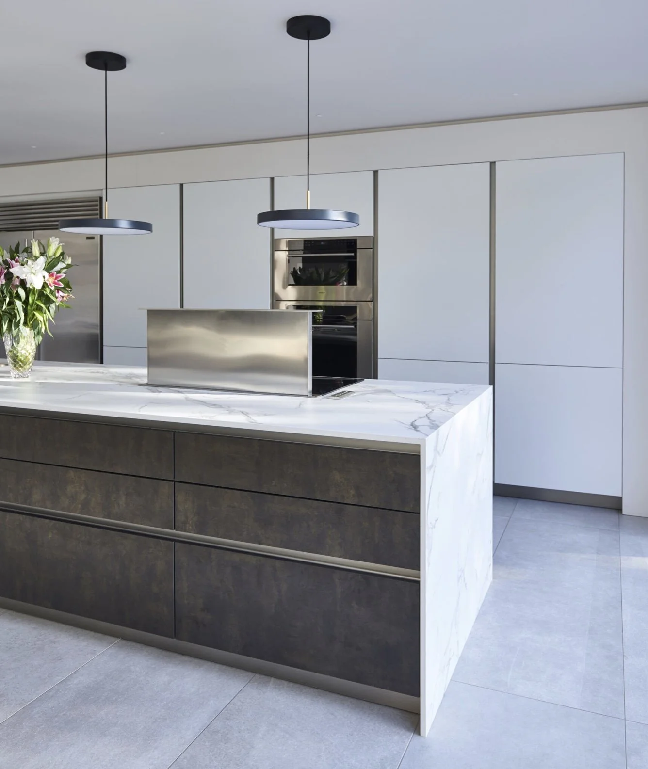 Modern kitchen with a marble island, black drawers, built-in stainless steel oven, and two black pendant lights.