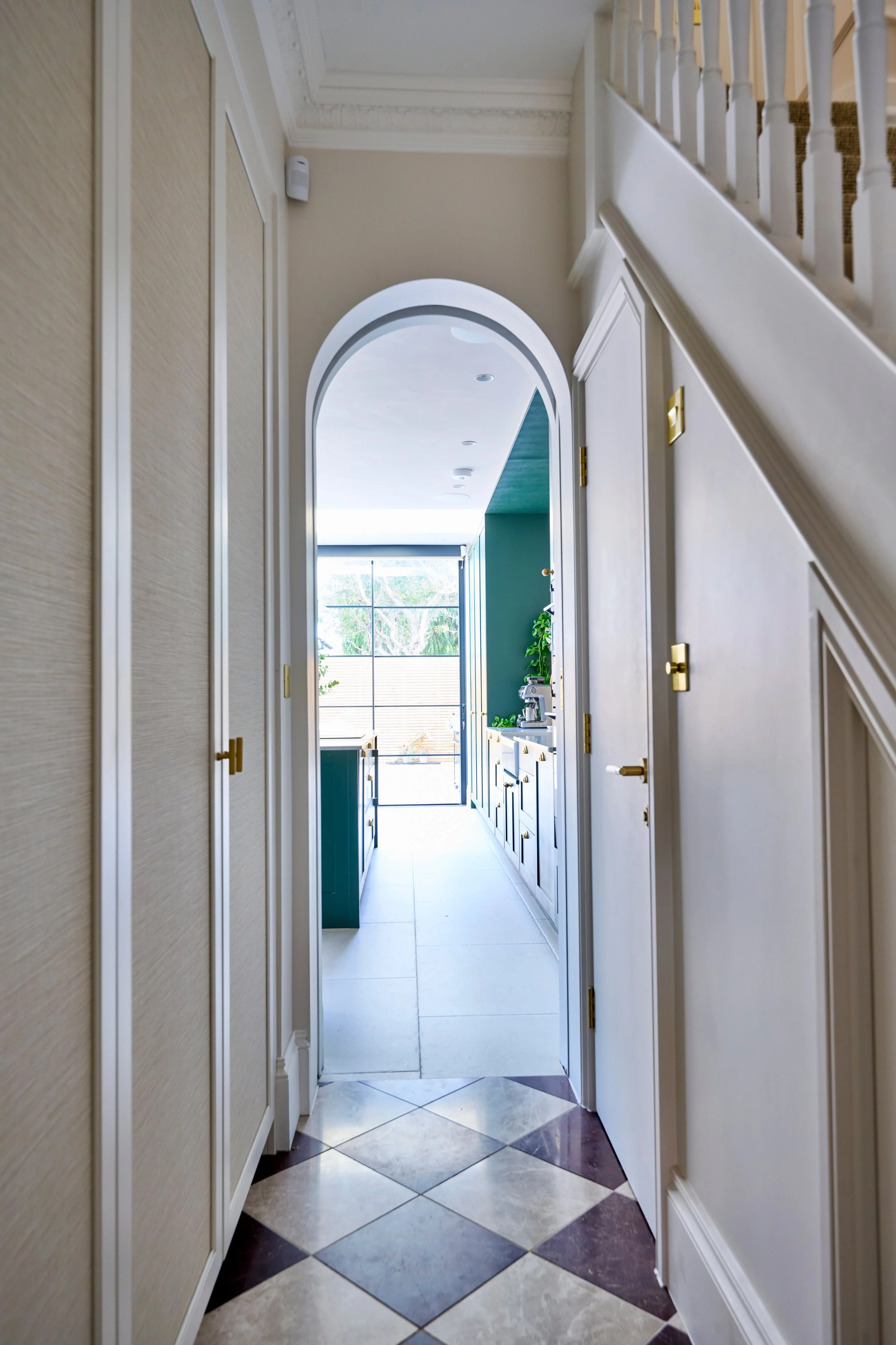 A narrow hallway leading to a room with a large window, blue walls, and a white ceiling. The hallway has beige walls, white trim, and a marbled floor with diamond patterns in beige, brown, and white. There is a staircase with a white railing and beig