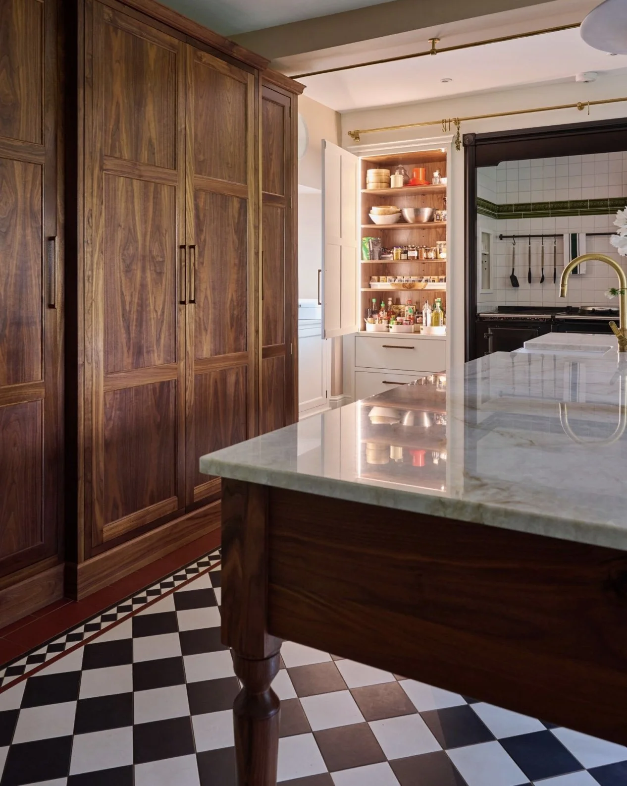 Kitchen with wooden cabinets, a marble countertop, and an open pantry with various food items. A door opens to the stove area with white tiled walls and green trim.
