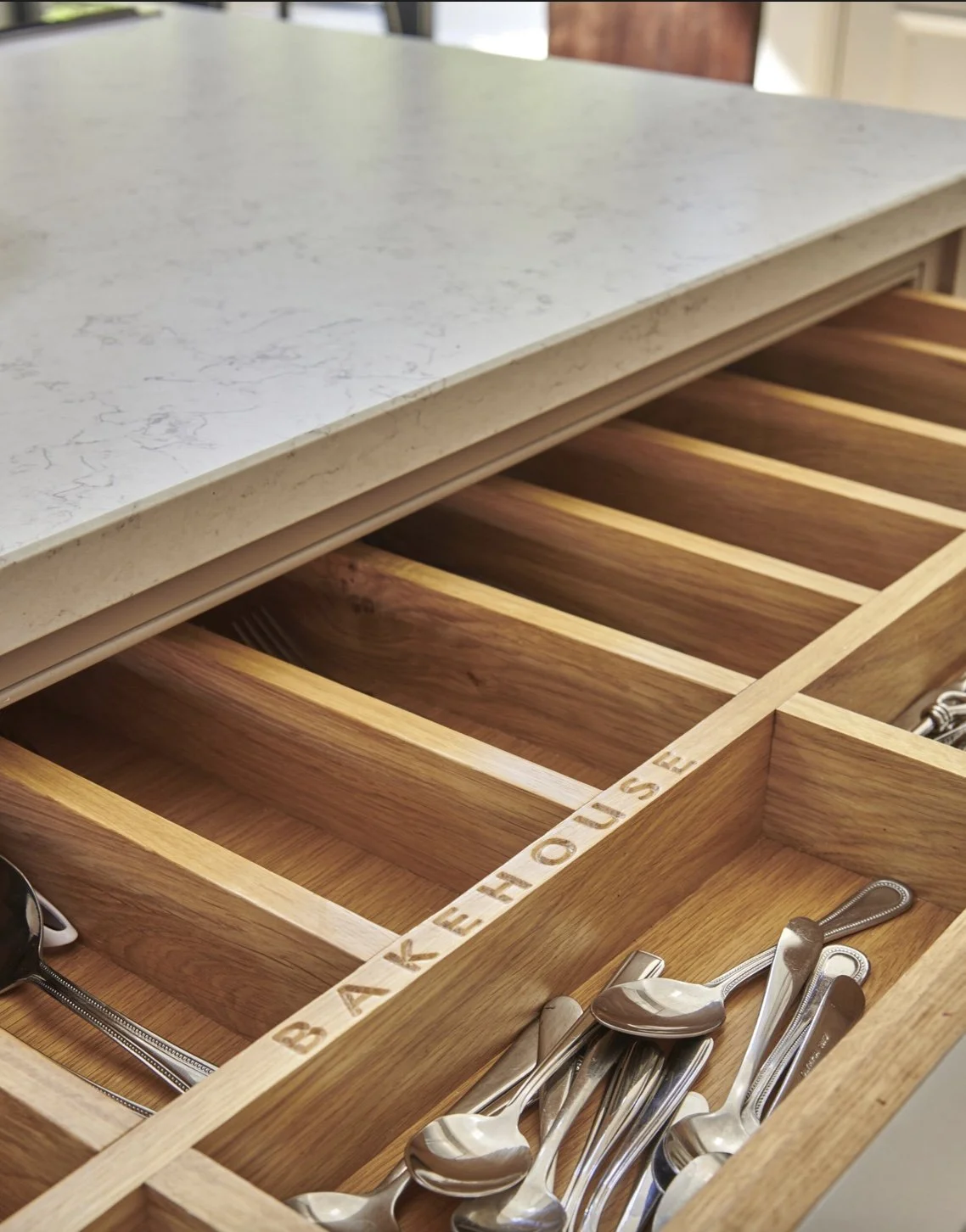 Wooden drawer organizer labeled 'BAKEHOUSE' with compartments for utensils, containing silverware, located under a white marble countertop.