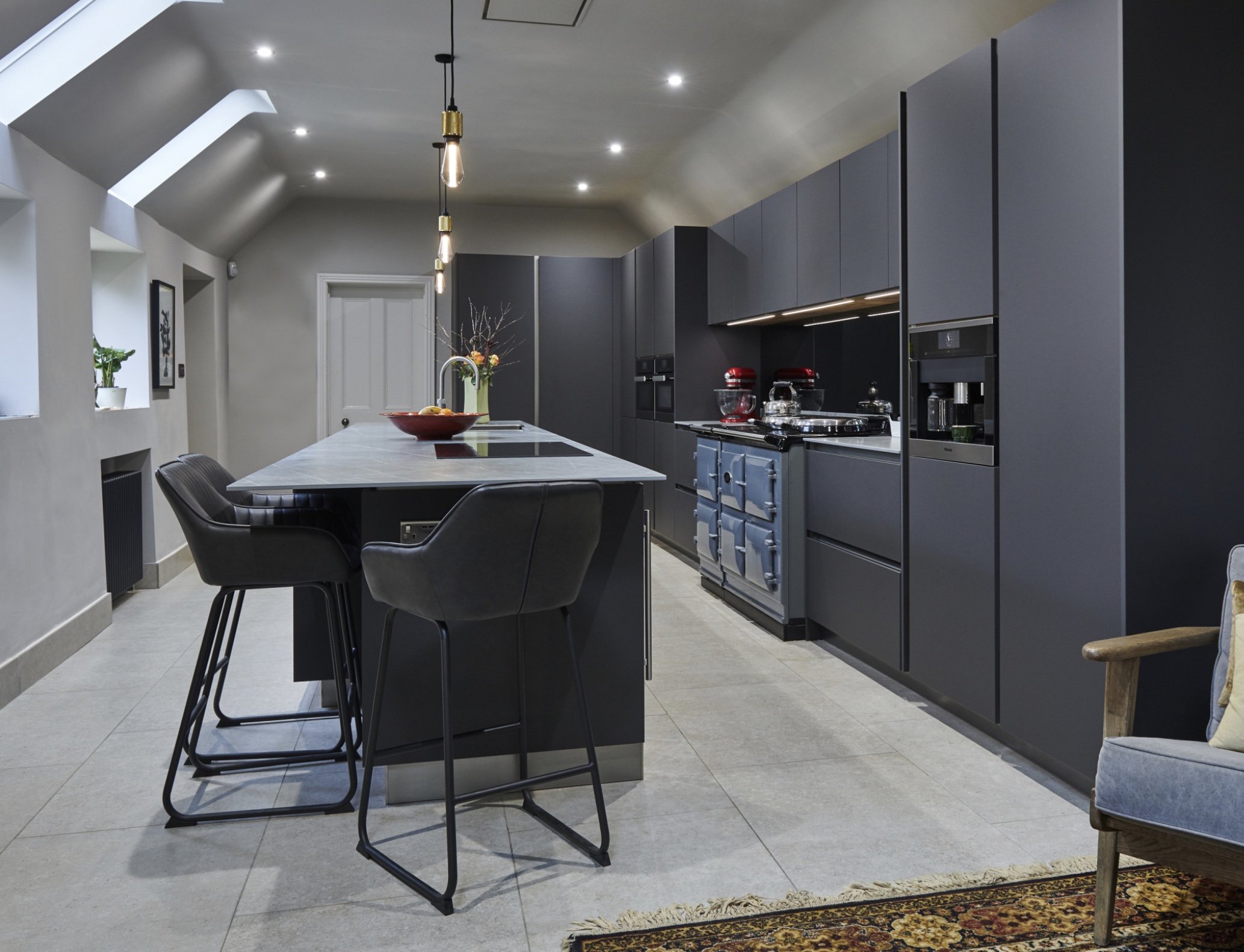 Modern kitchen with dark cabinets, a large island with a marble countertop, black chairs, stainless-steel appliances, and pendant lighting.