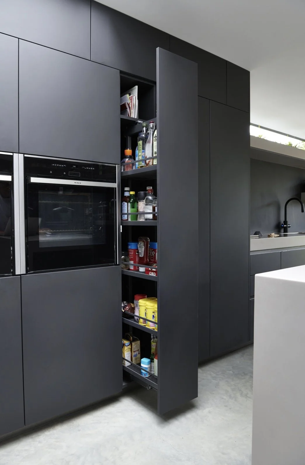 A modern kitchen with dark gray cabinets and built-in oven, featuring an open pull-out pantry with various condiments, spices, and pantry items.