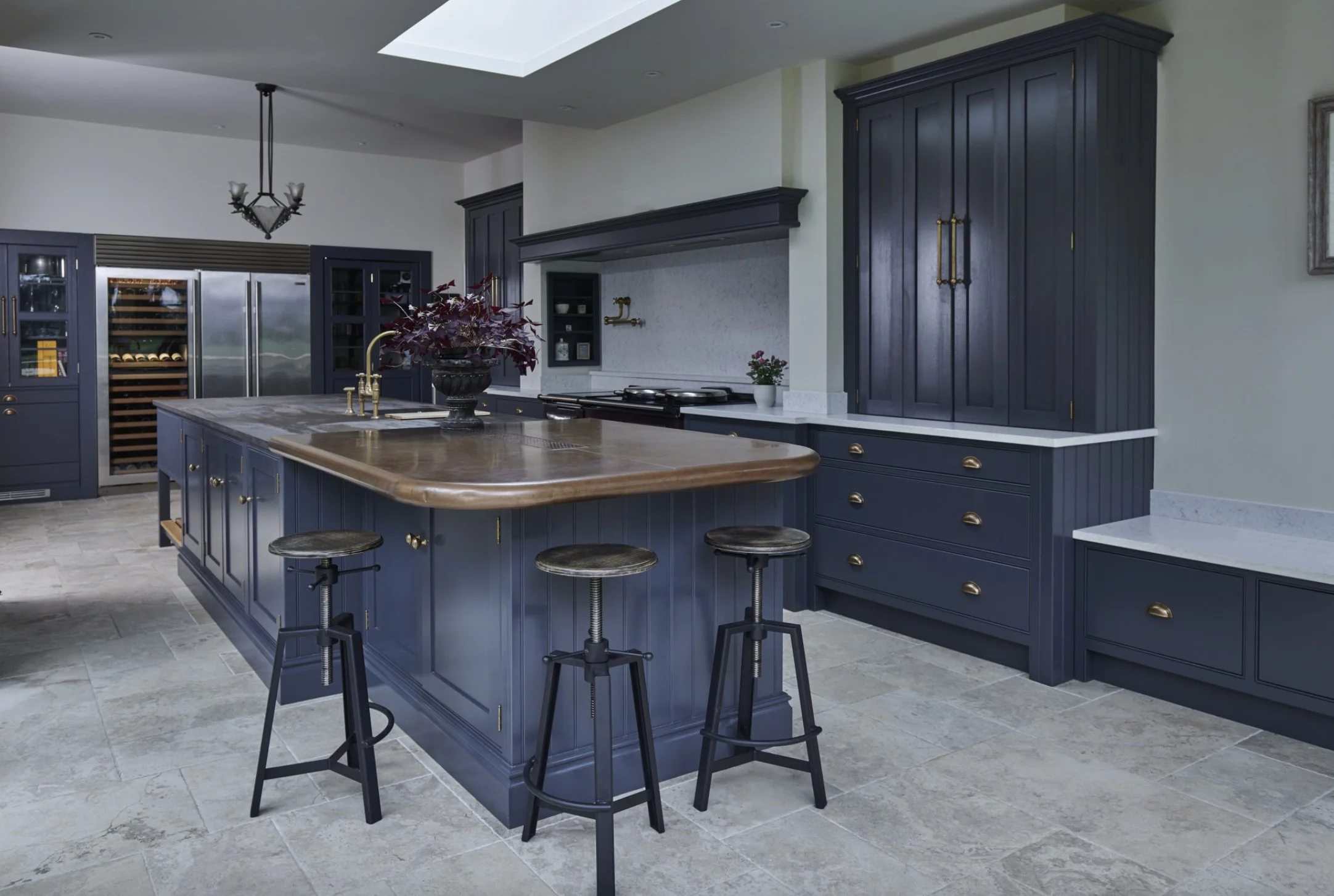 Modern kitchen with navy blue cabinets, a large island with a wooden countertop, black bar stools, and stainless steel appliances in the background.