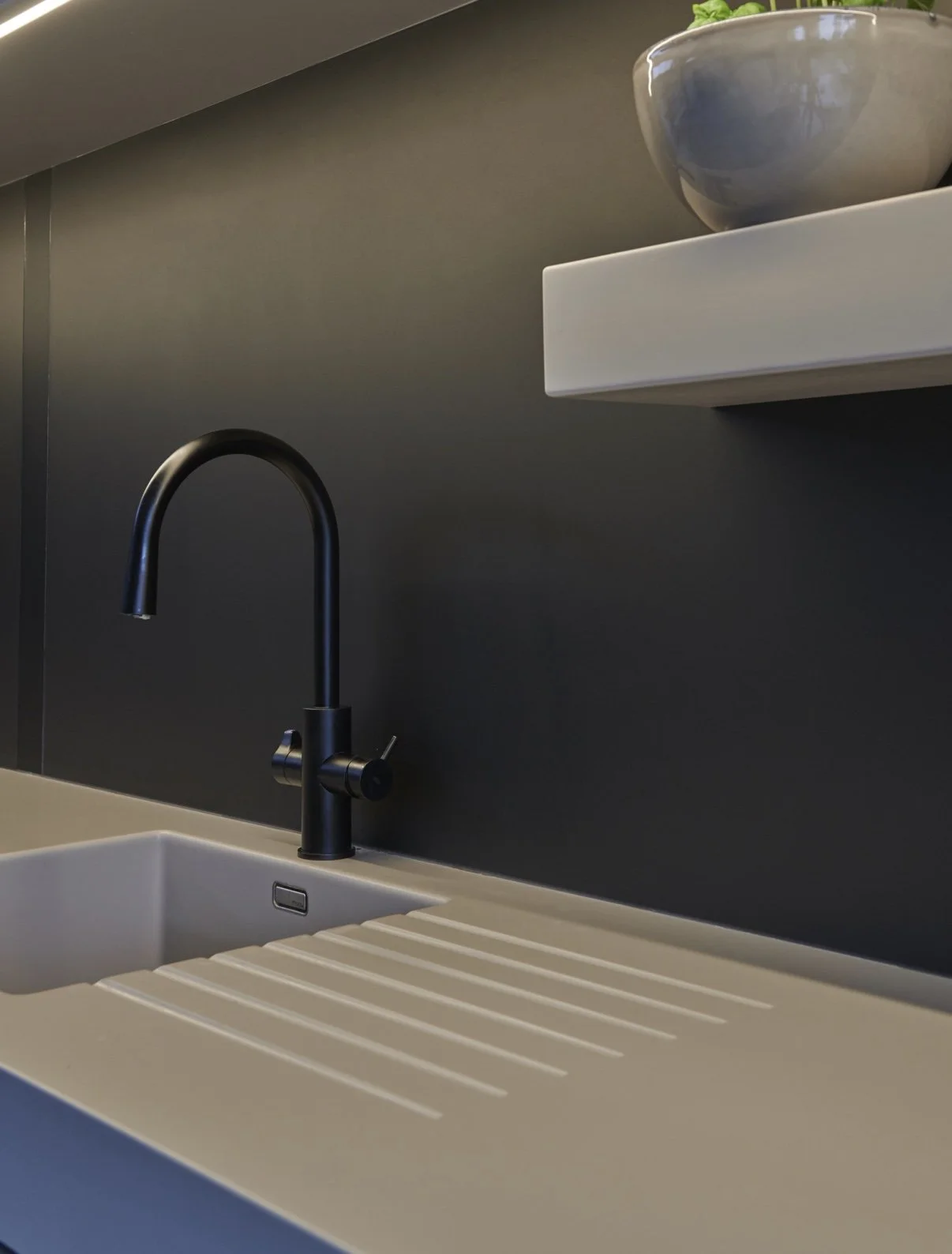 Close-up of a modern kitchen sink with a black faucet and a dark grey wall in the background, with a white shelf and a grey bowl on top.
