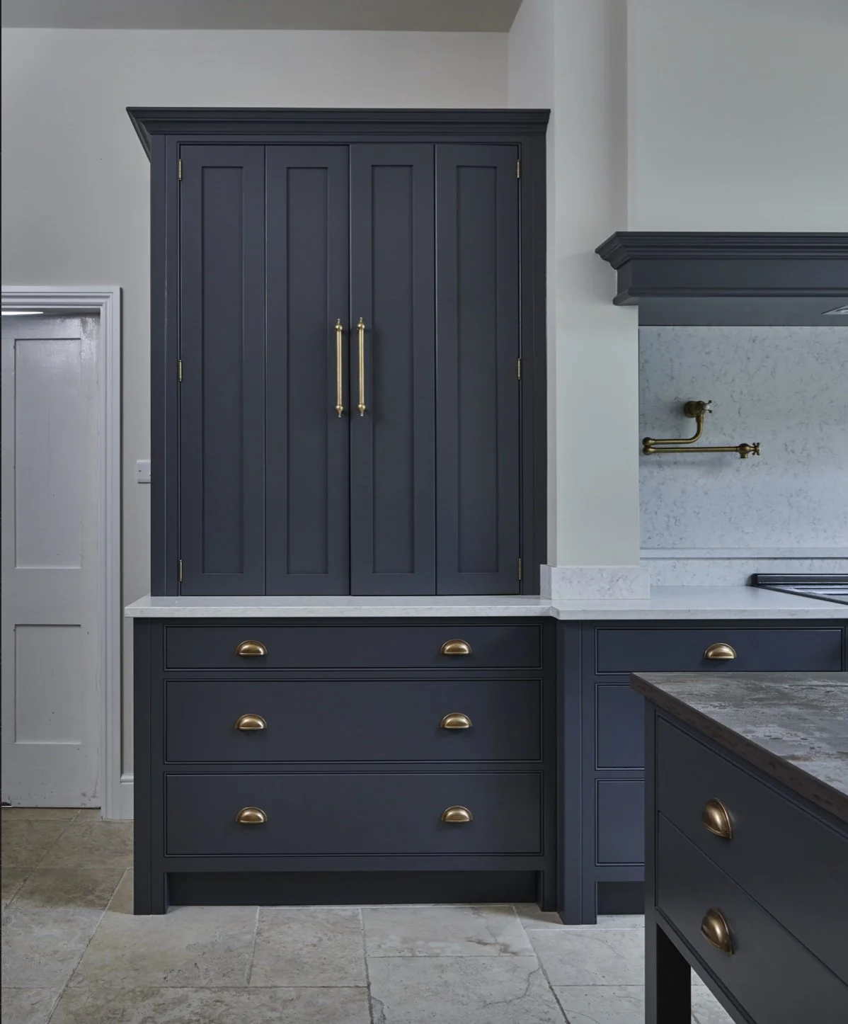 Interior kitchen with dark blue cabinetry, white marble countertops, and gold hardware, including cabinet handles and faucet, on the wall.
