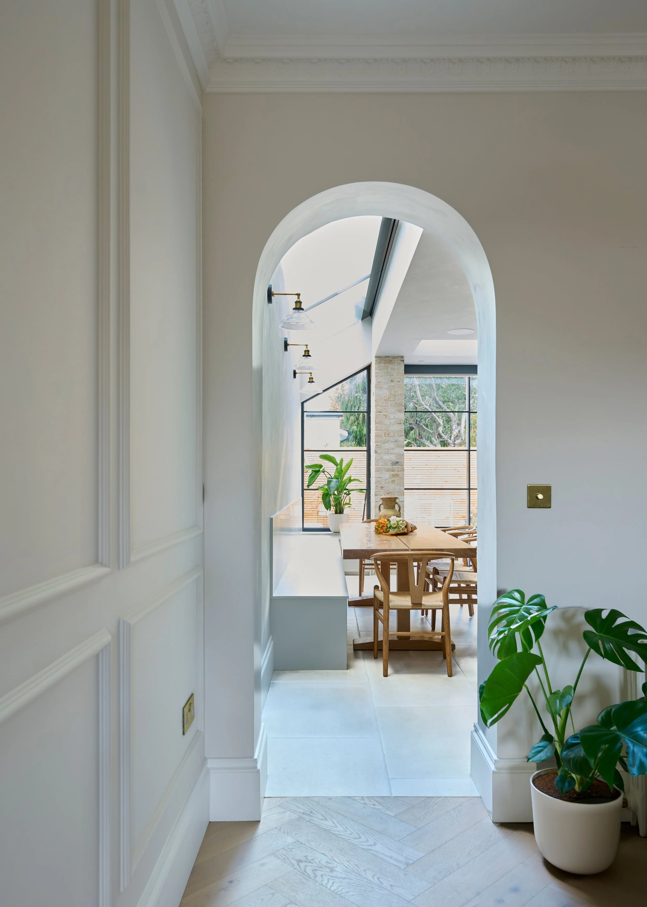 A bright dining area viewed through an arched doorway, with a wooden table, chairs, and potted plants near large windows.