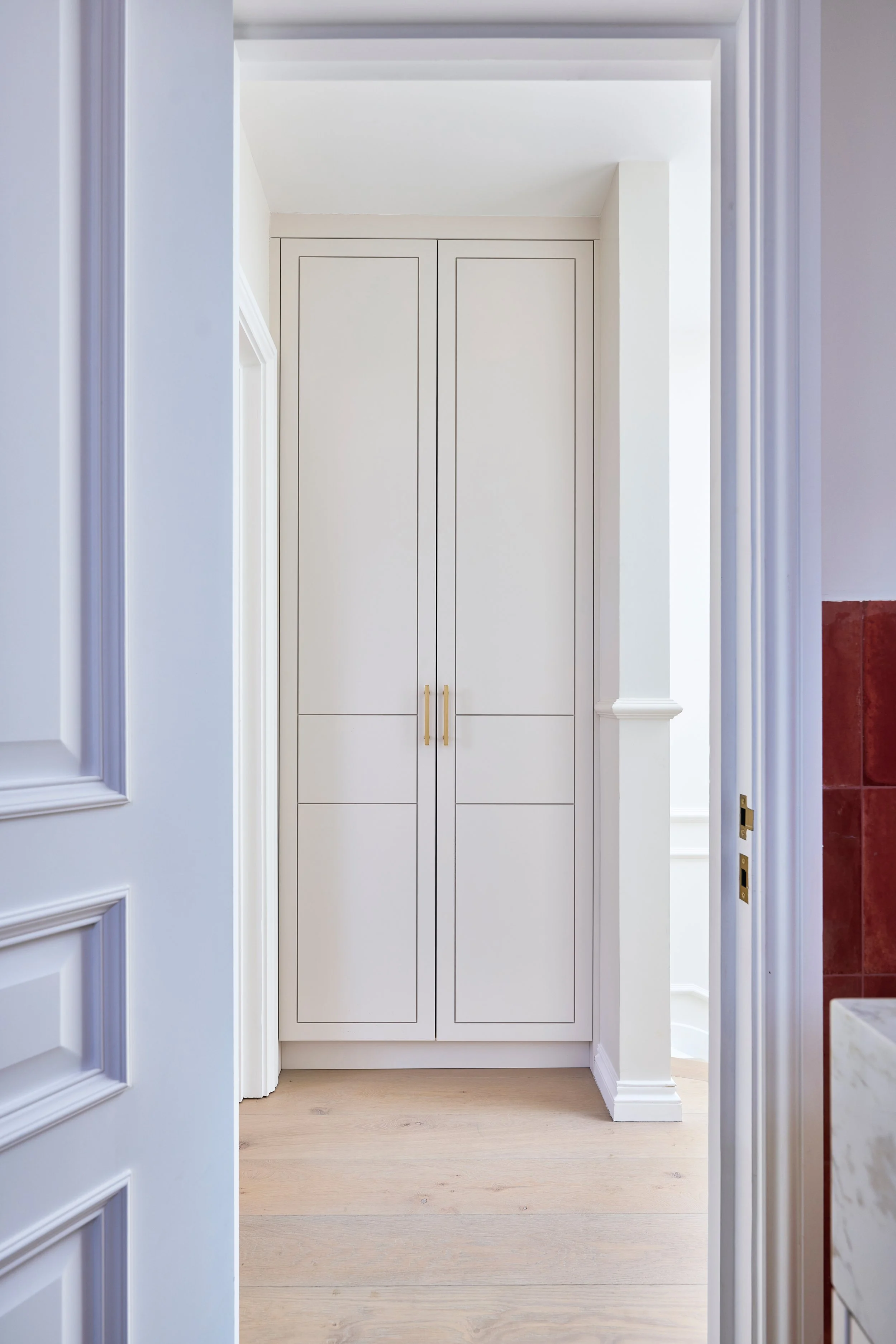View through an open door revealing a white built-in closet with two doors and brass handles in a bright room with white walls and light wood flooring.
