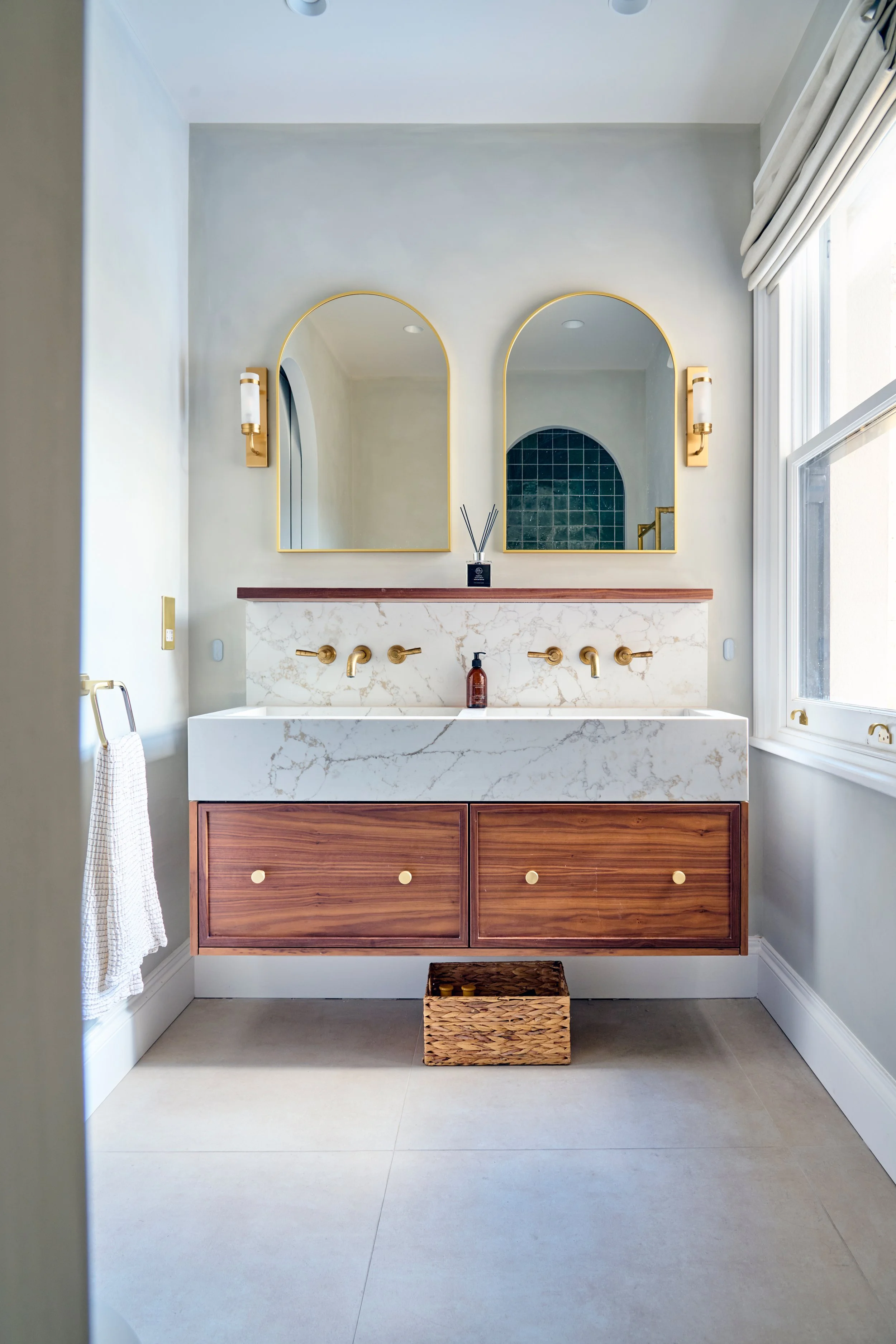 Modern bathroom vanity with two gold mirrors and matching wall sconces, a marble countertop with a vessel sink, gold fixtures, a wooden cabinet, and a wicker basket on the floor.