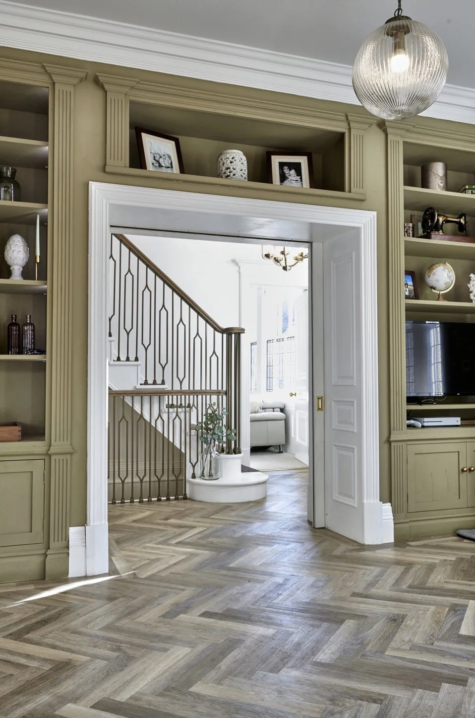Interior view of a living room with built-in shelves, a staircase with a metal railing, decorative items, framed photos, and a bright sitting area in the background.