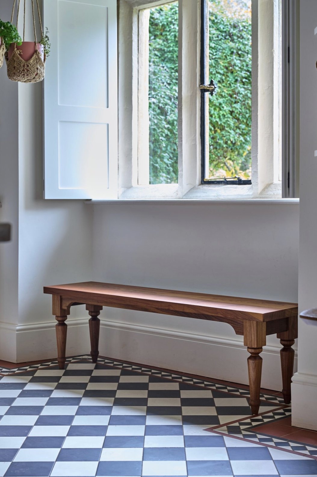 Interior of a room with a large open window showing greenery outside, a wooden bench against the wall, and a patterned tile floor, with hanging plants on the left.