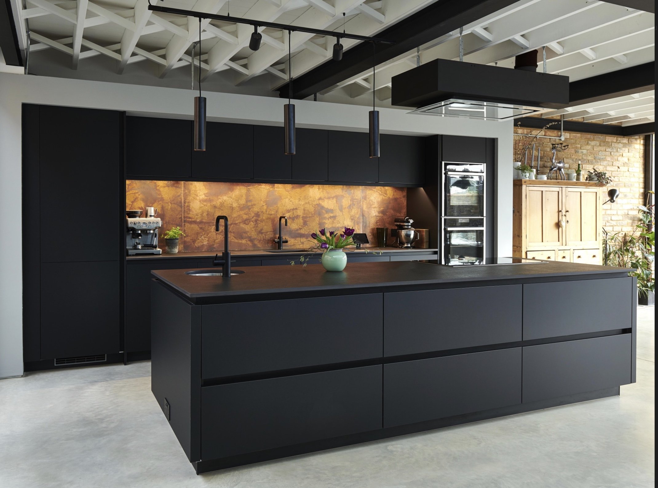 Modern kitchen with black cabinetry and island, copper backsplash, and wooden storage cabinet, decorated with plants and kitchen appliances.