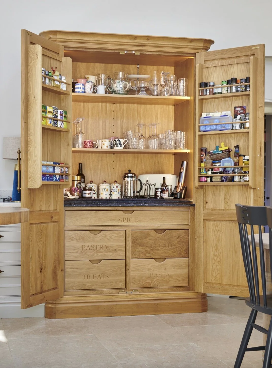 Open wooden kitchen cabinet with labeled drawers for spices, pastry, treats, bread, and pasta, containing glassware, mugs, jars, and various pantry items.