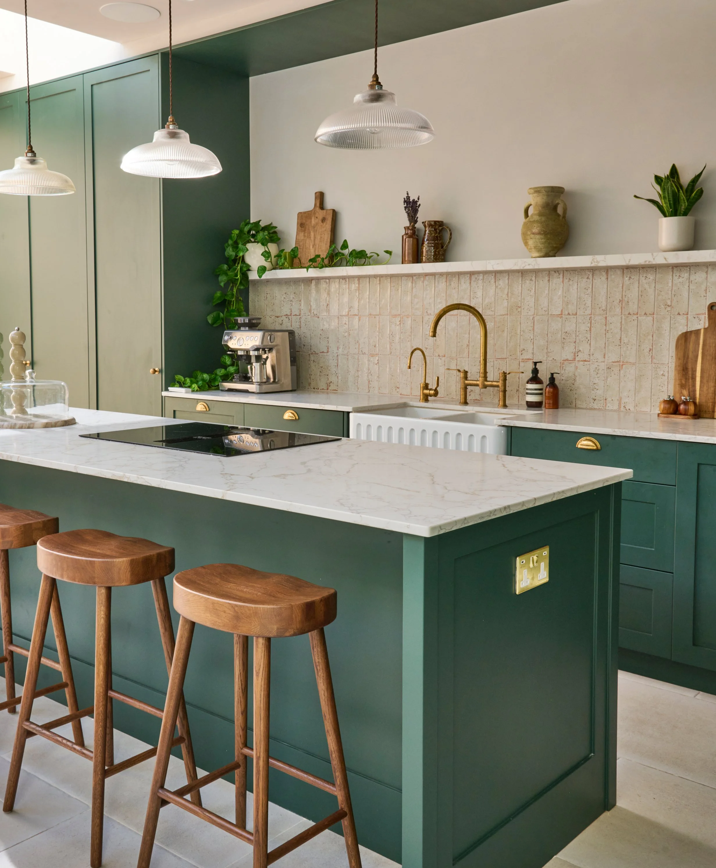 A modern kitchen with a green island and matching cabinets, white marble countertop, hanging pendant lights, and decorative vases and plants on the shelf, with three wooden bar stools in front.