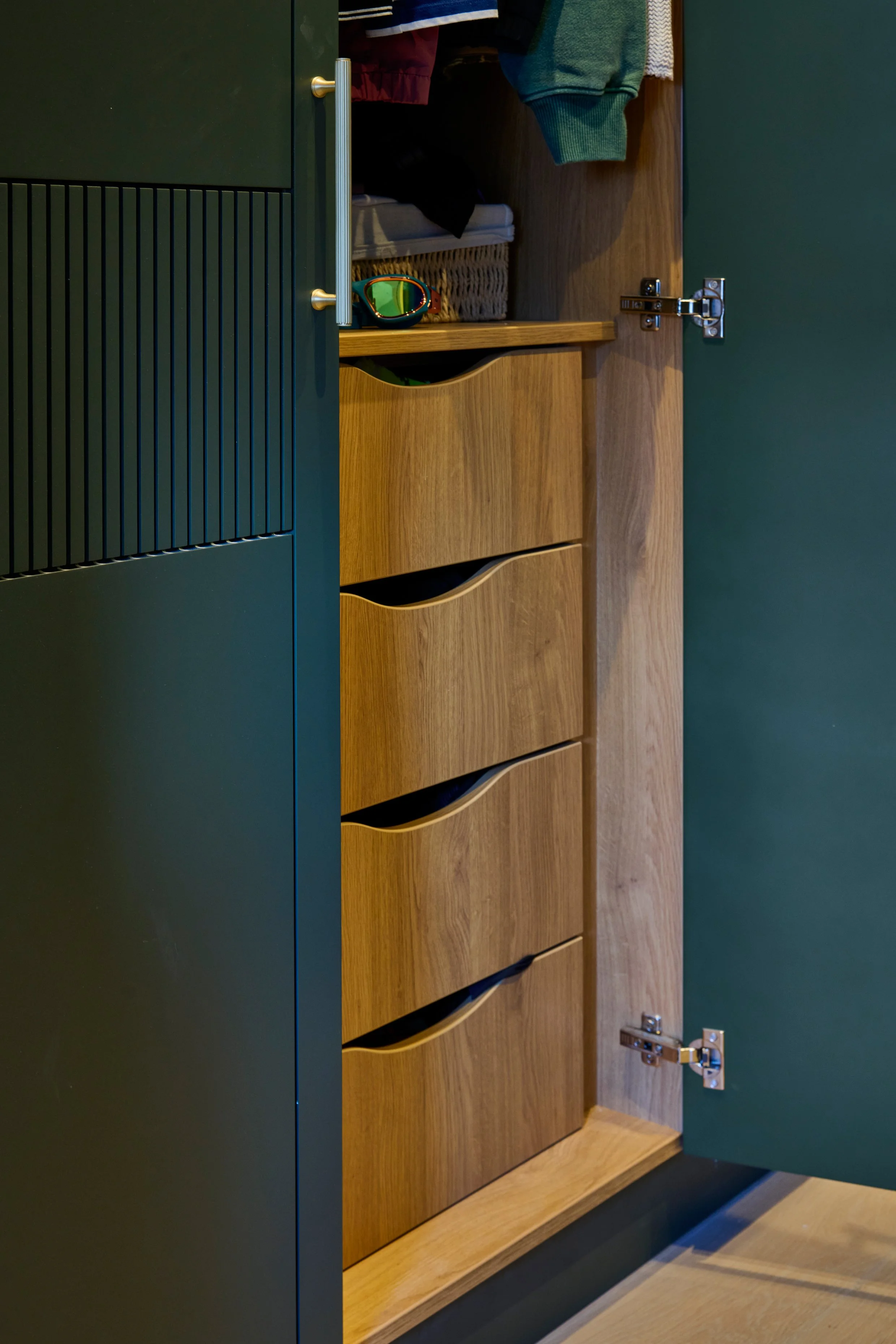 A partially open wooden cabinet with drawers and a hanging rack inside, holding clothes, and a pair of ski goggles resting on top.