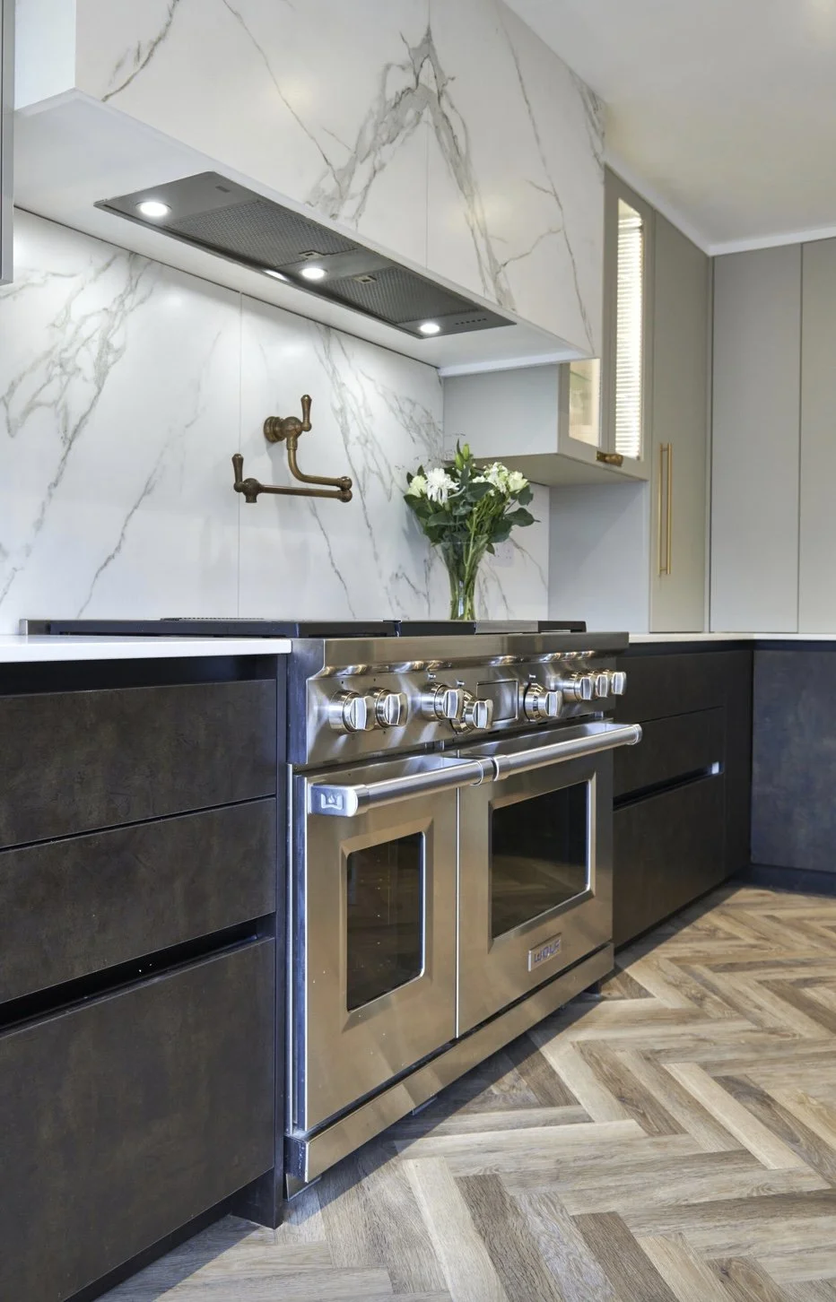 Modern kitchen with marble backsplash, brass faucet fixtures, a stainless steel stove and oven, dark cabinetry, a vase with white flowers, and wood-patterned flooring.