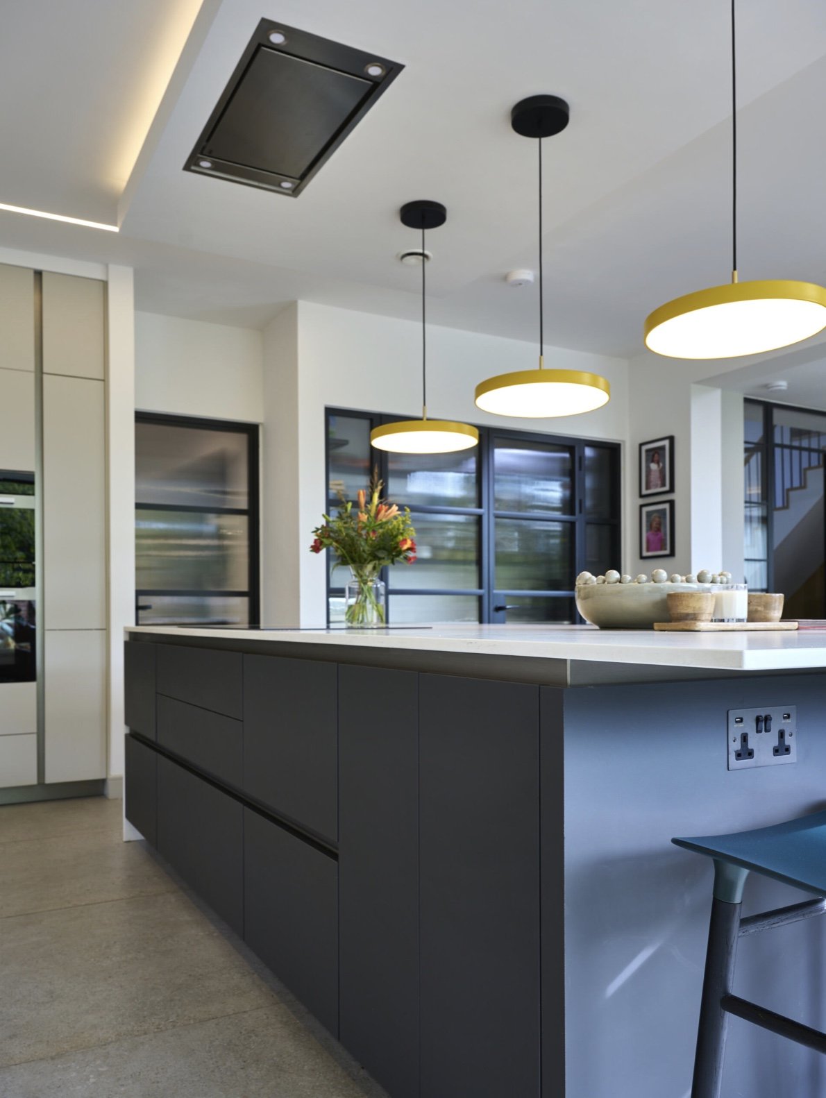 Modern kitchen with black cabinets, white countertops, three yellow pendant lights, a vase of colorful flowers, and black-framed windows.
