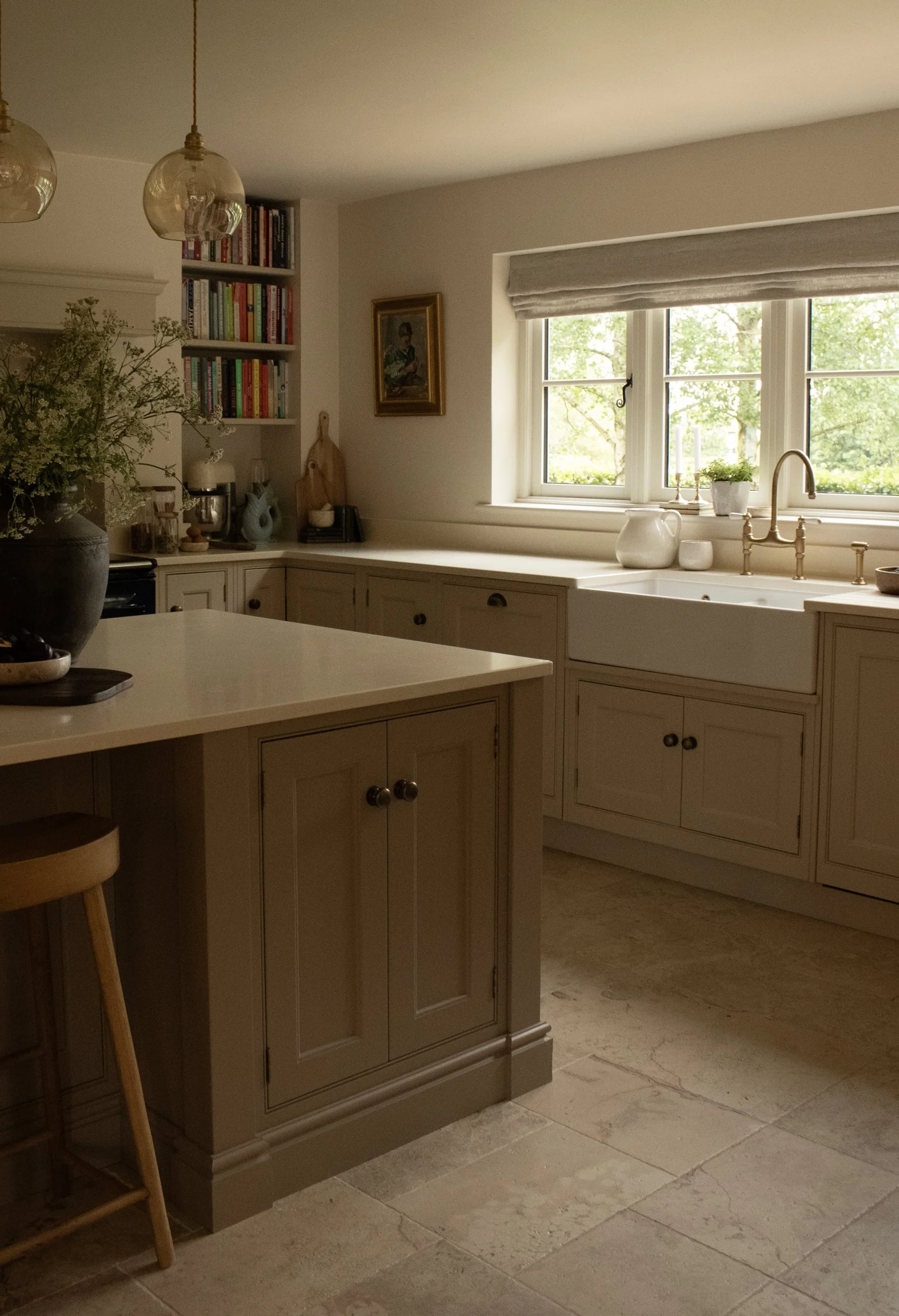 A cozy kitchen with a large window overlooking green trees. The kitchen features beige cabinetry, a white countertop, and decorative items like pottery and a painting on the wall. There are two glass pendant lights hanging from the ceiling and a plan
