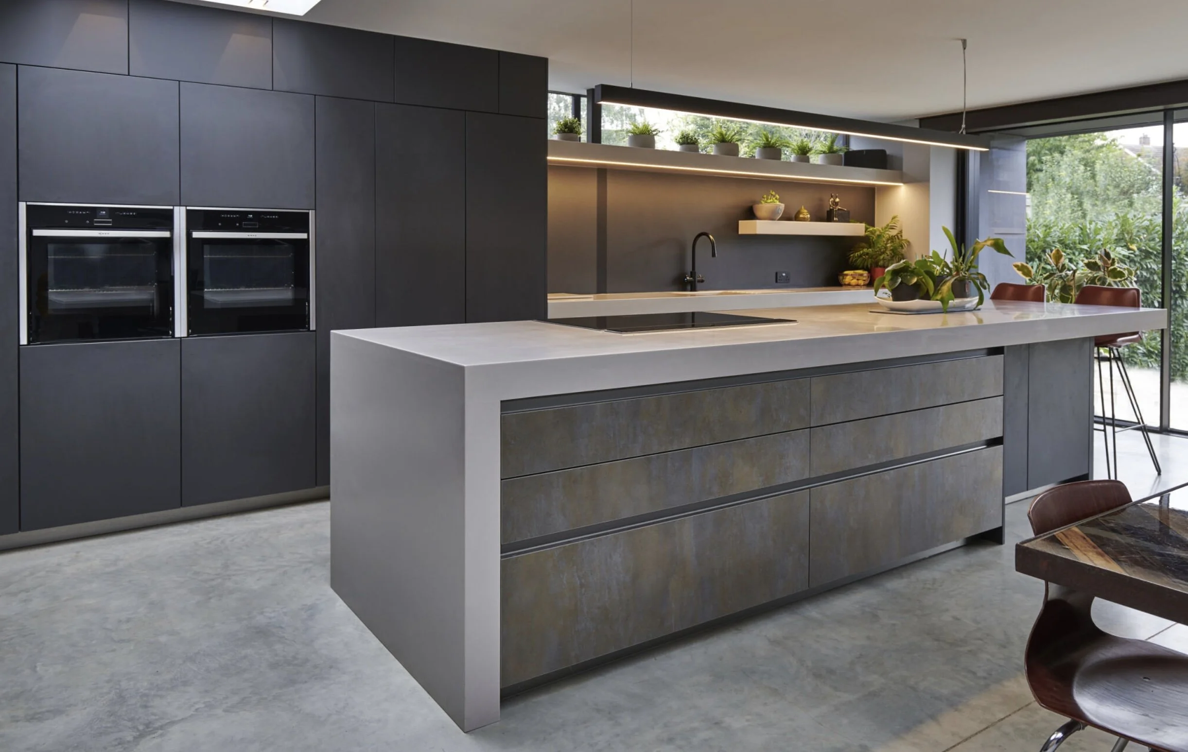 Modern kitchen with dark cabinets, a large island with a white countertop and drawers, black appliances, green plants on shelves and outside, and red chairs around a dining table.
