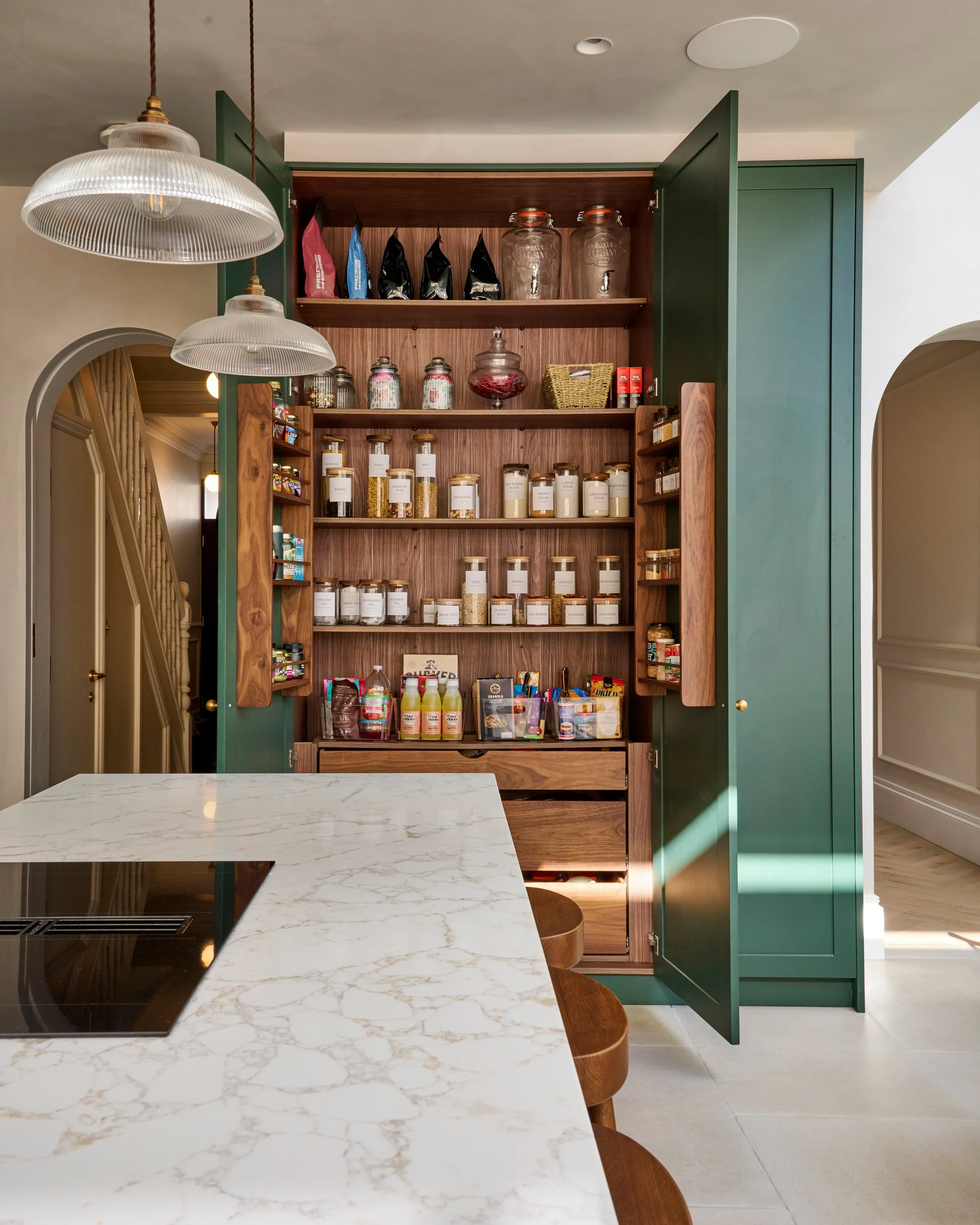 Open green kitchen cabinet revealing shelves with jars of spices, dried herbs, and snacks, alongside decorative glass jars and a basket, with a marble countertop in the foreground.