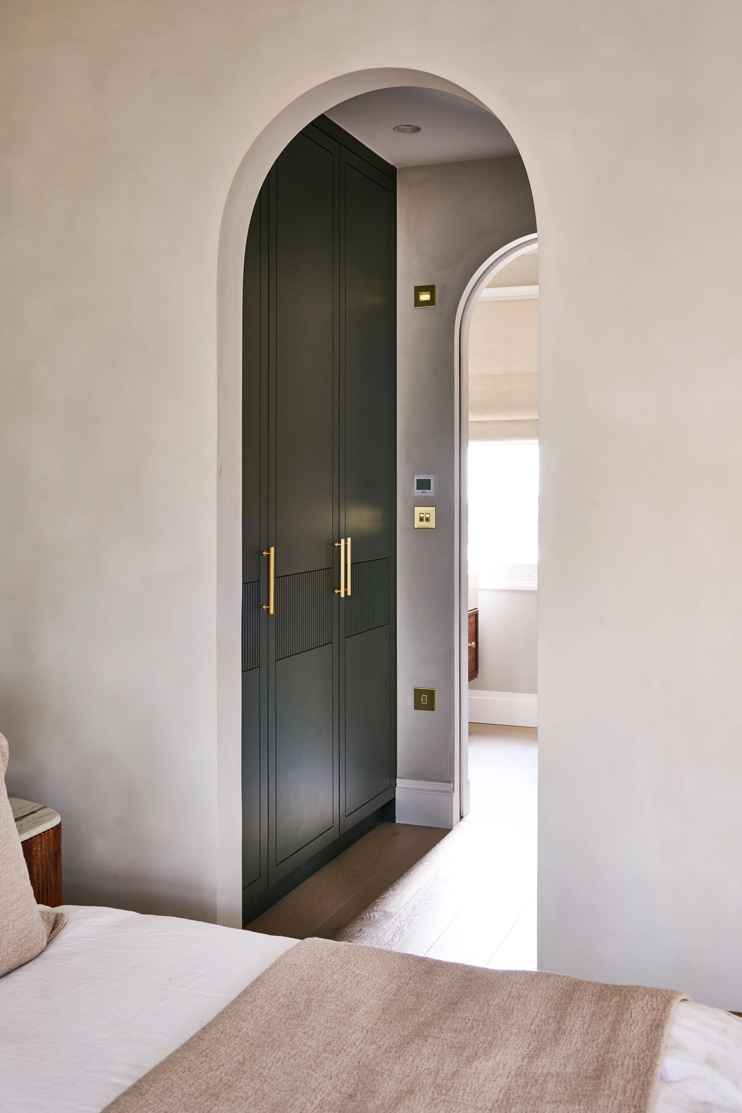 A bedroom with a view through an arched doorway, featuring a large green wardrobe with gold handles, and a portion of a bed with beige linens in the foreground.