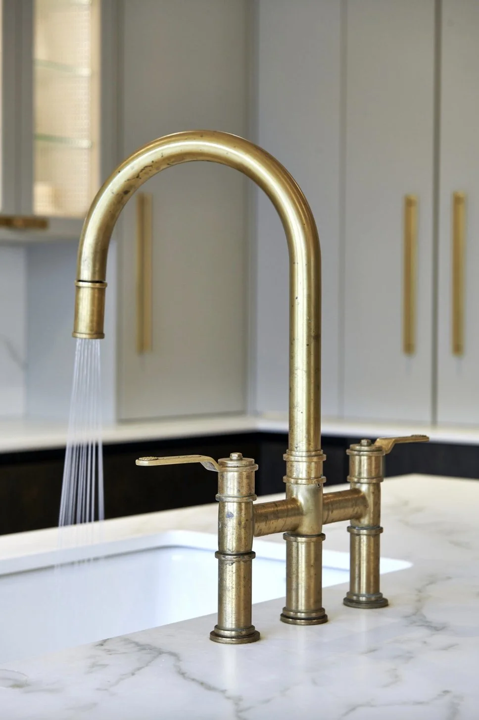 Gold kitchen faucet with water running, mounted on a white marble countertop in a modern kitchen with light cabinets in the background.
