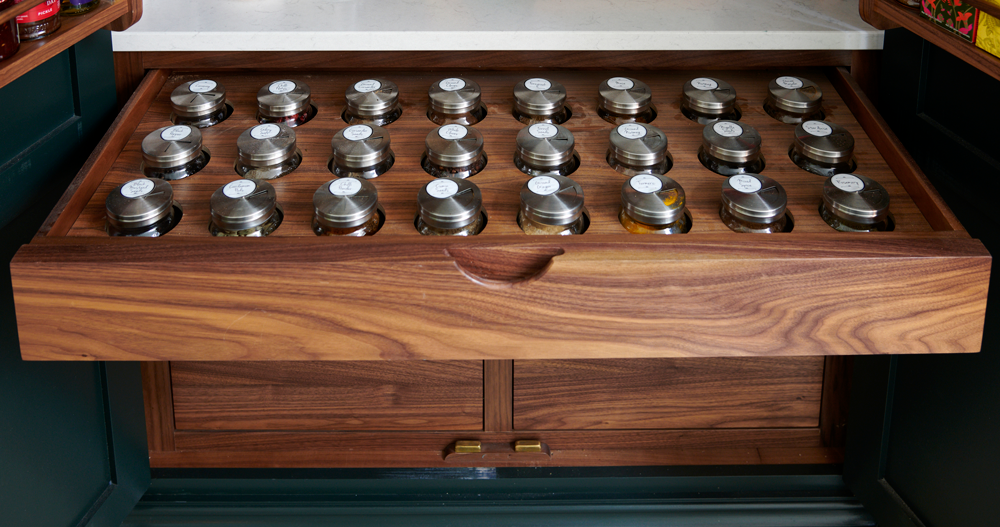 Display of various spices in small glass jars with metal lids, arranged on a wooden shelf in a spice shop.