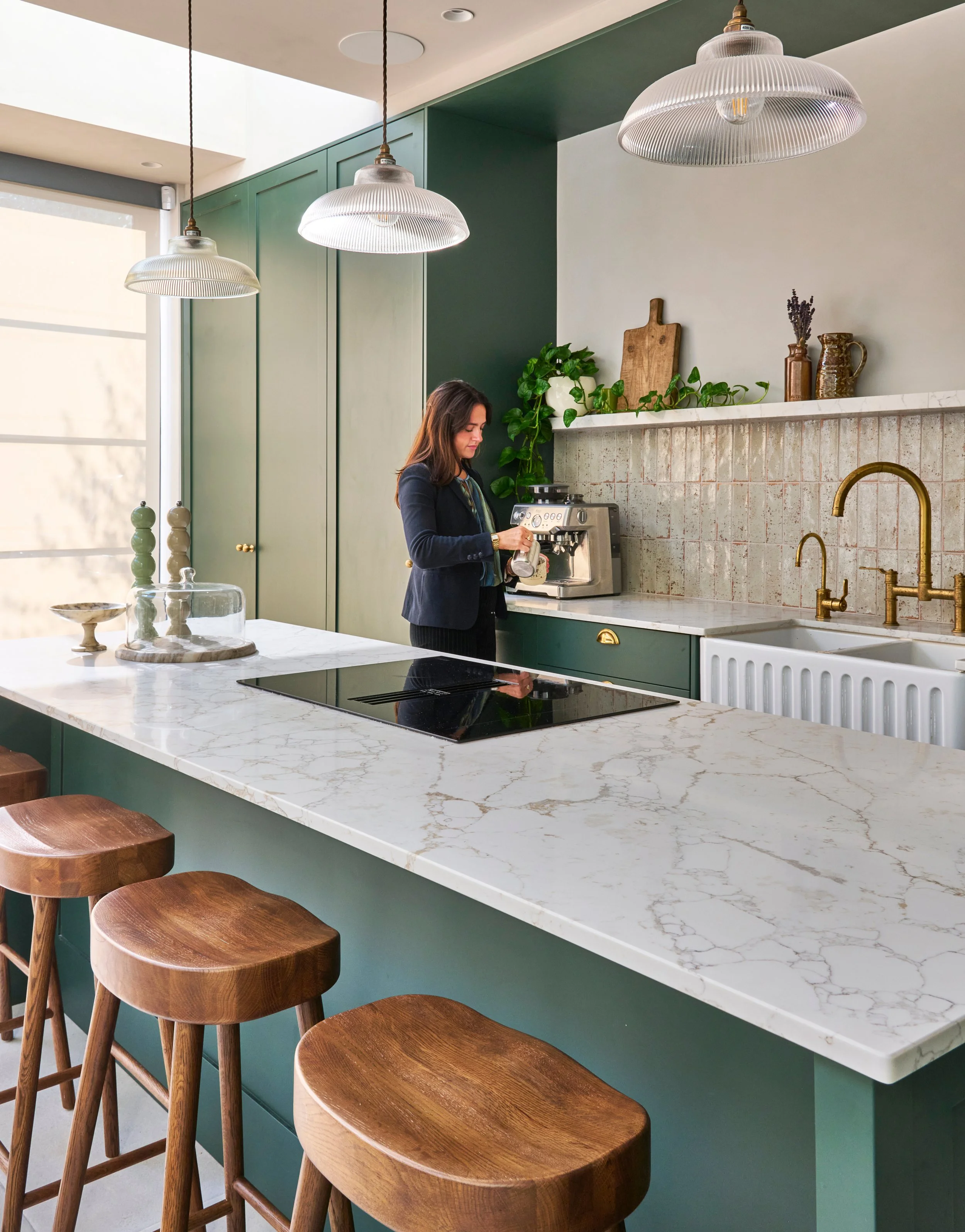 A woman in a dark blazer stands at a kitchen counter with a coffee machine. The kitchen has green cabinets with gold hardware, a marble countertop, and brass faucets. Hanging pendant lights with ribbed glass shades illuminate the space. Bar stools wi