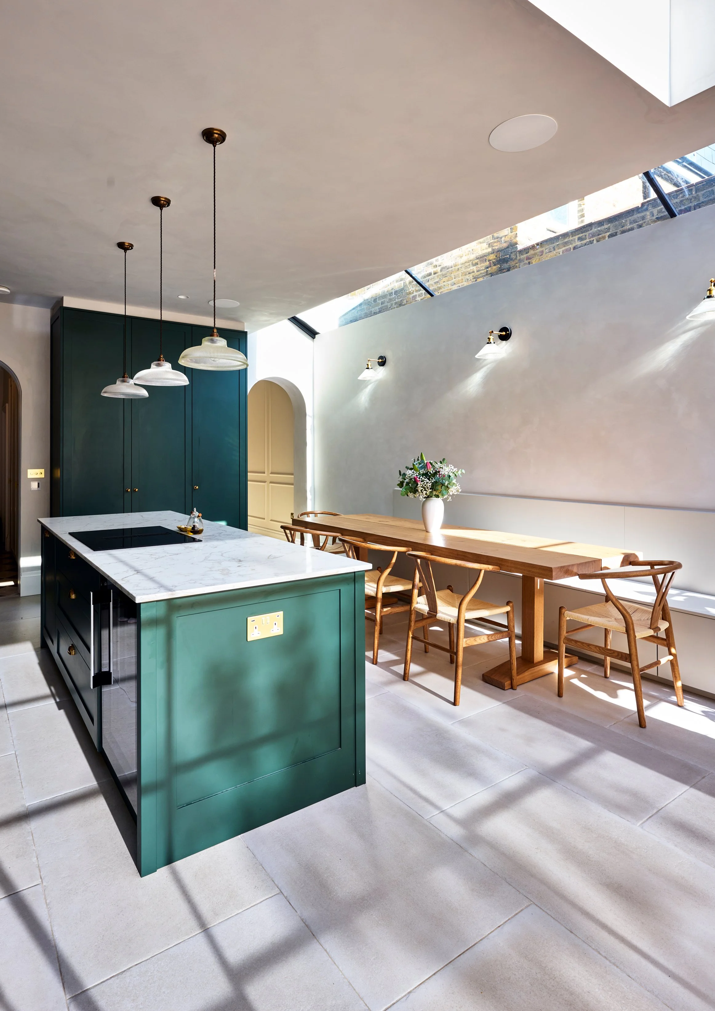 Modern kitchen with a white marble island, teal cabinets, and a wooden dining table with chairs. There are hanging pendant lights and wall sconces, and a vase of flowers on the table. Skylights and brick walls provide natural light.