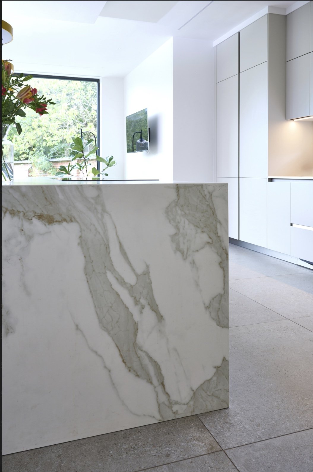 Modern kitchen with white cabinets, a marble island, a window with greenery outside, and a wall-mounted TV.