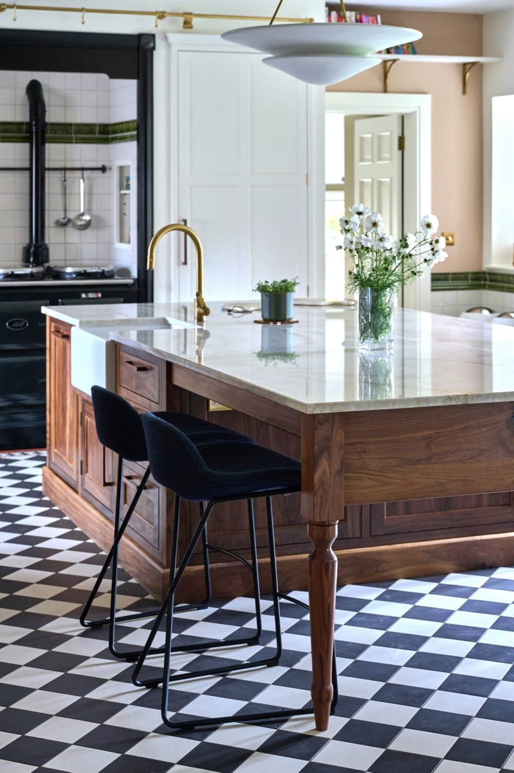 A modern kitchen with a wooden island countertop, black bar stools, a black stove with a chimney, a white cabinet, a vase of white flowers, a small potted plant, and black and white checkered floor tiles.