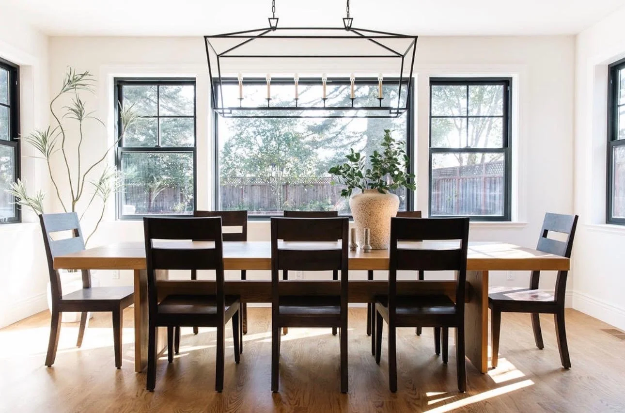 Dining room with wooden table, eight black chairs, a large vase with greenery, and a modern chandelier, with large windows showing a backyard.