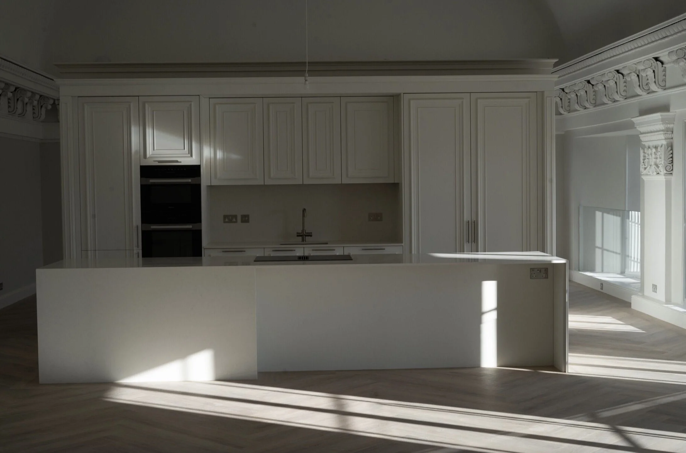 Empty kitchen with white cabinets, a white island, and sunlight streaming through windows, casting shadows on the wooden floor.