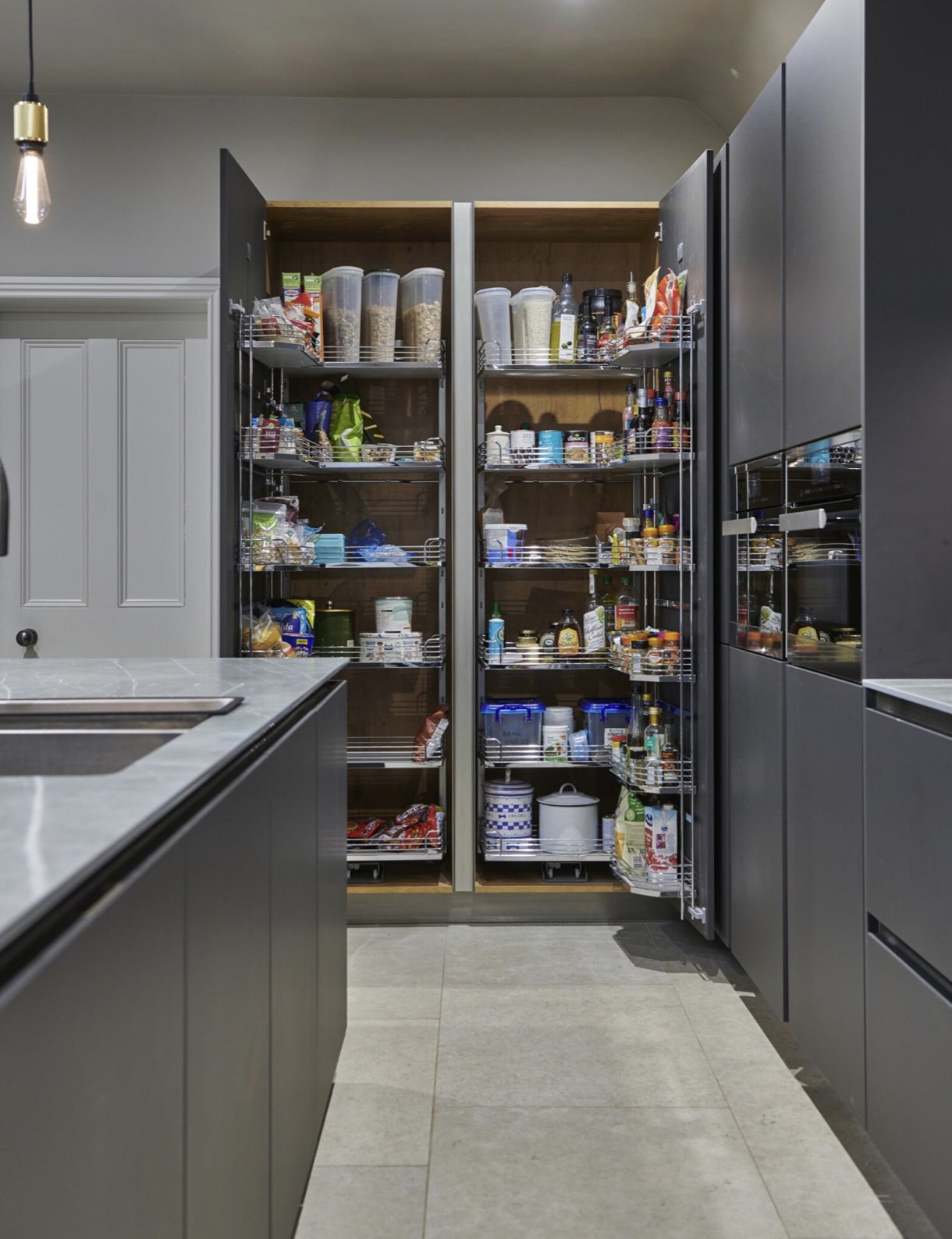 Kitchen pantry with open shelves filled with various food items, containers, and bottles, located between black cabinets and a gray countertop.