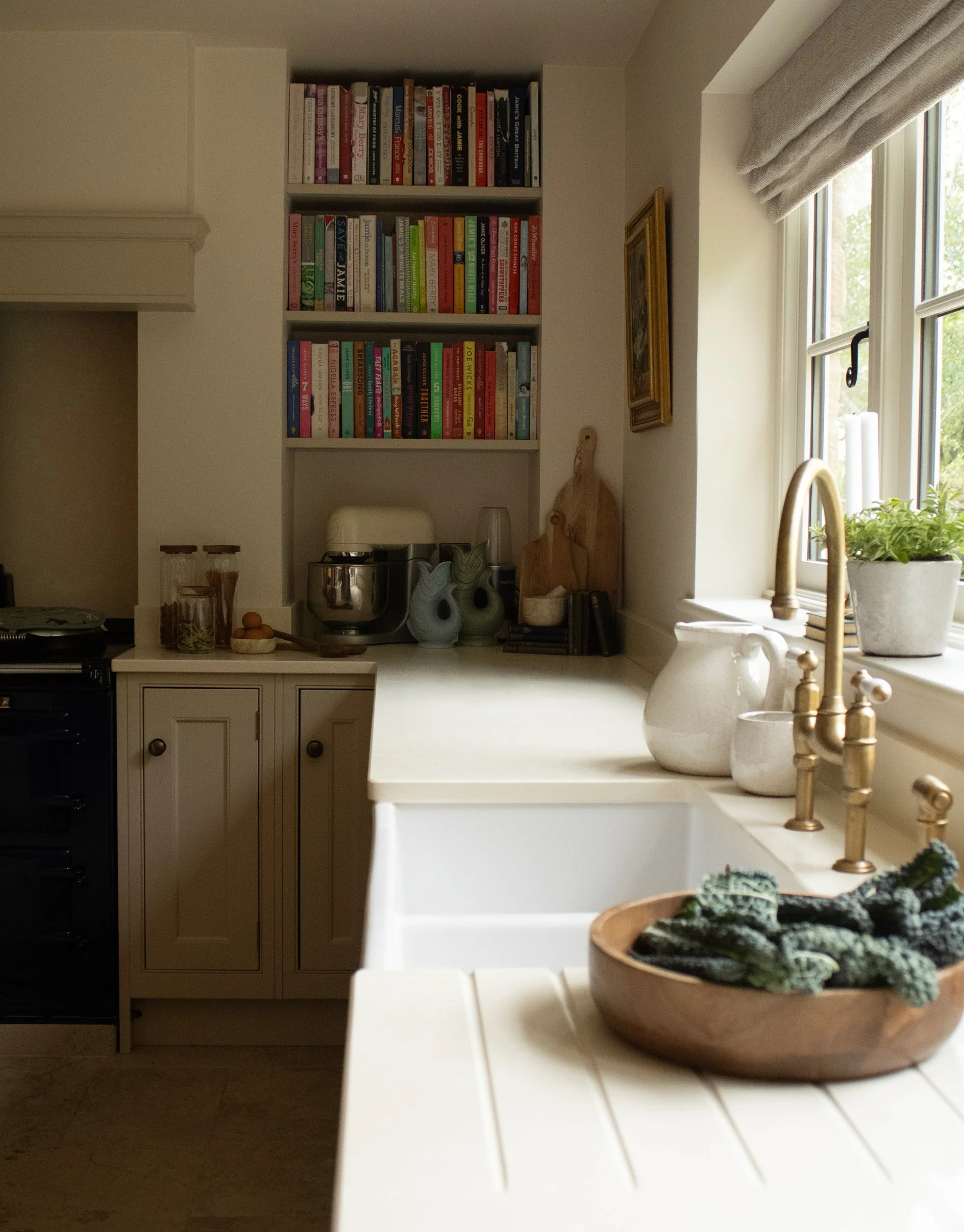 A kitchen with a white countertop, a window with curtains, a sink with a brass faucet, decorative vases, and a bowl with plants.