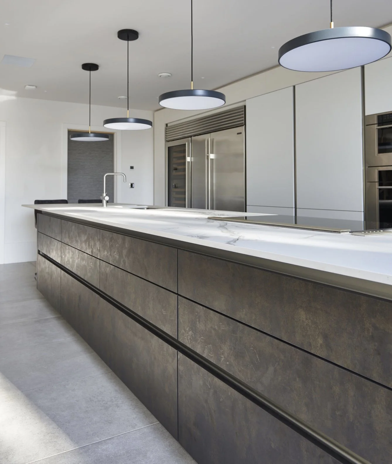 Modern kitchen with a large island featuring a marble countertop and dark textured drawers, pendant lights hanging from the ceiling, and stainless steel appliances including a refrigerator and built-in ovens.