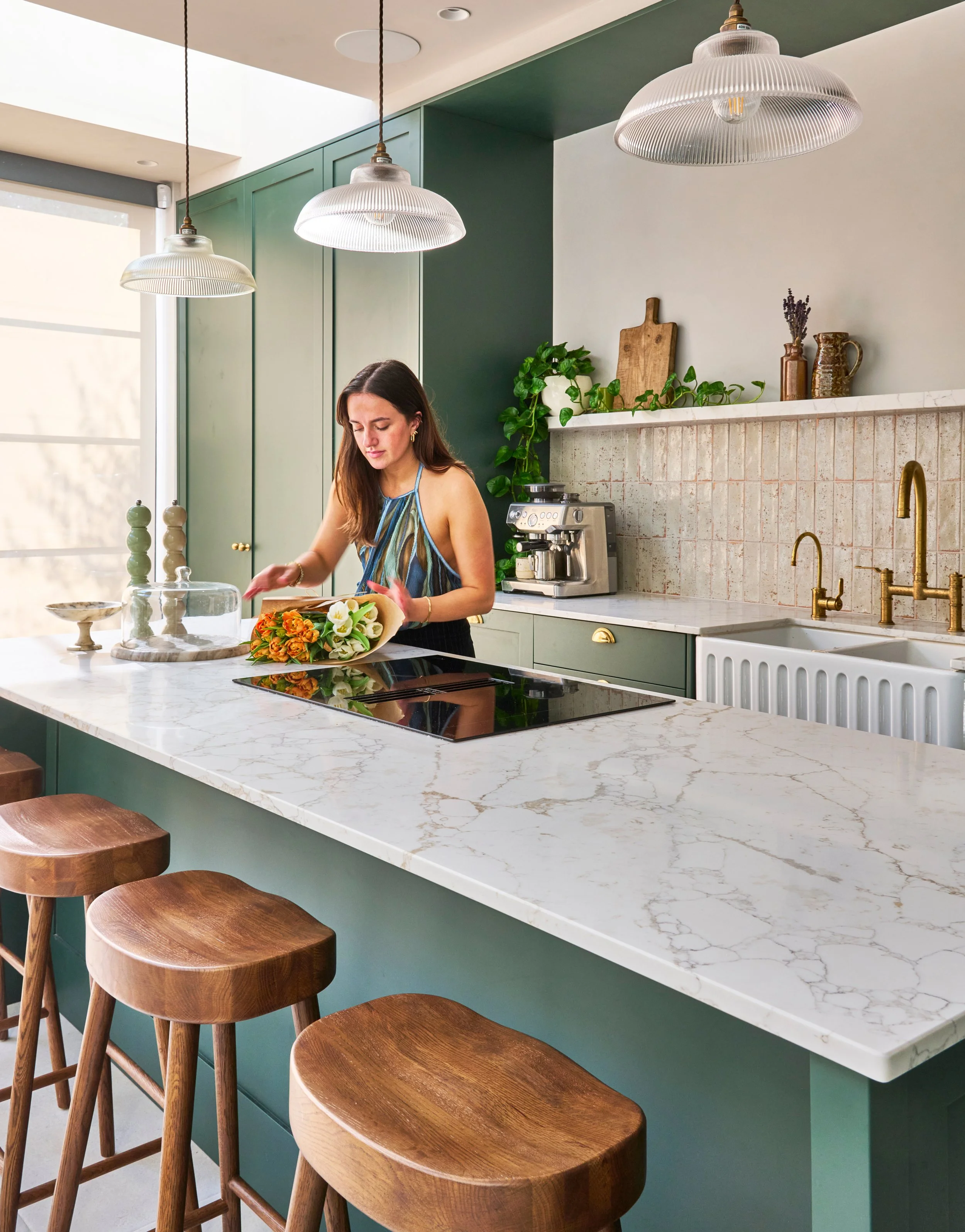 A woman in a colorful apron preparing food in a modern kitchen with green cabinets, a marble countertop, and wooden stools.
