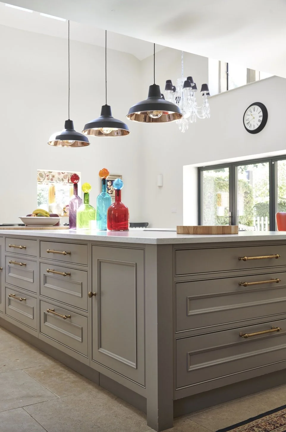Bright kitchen with gray cabinetry, colorful glass bottles on white countertop, large window, black pendant lights, and a chandelier, with a wall clock.