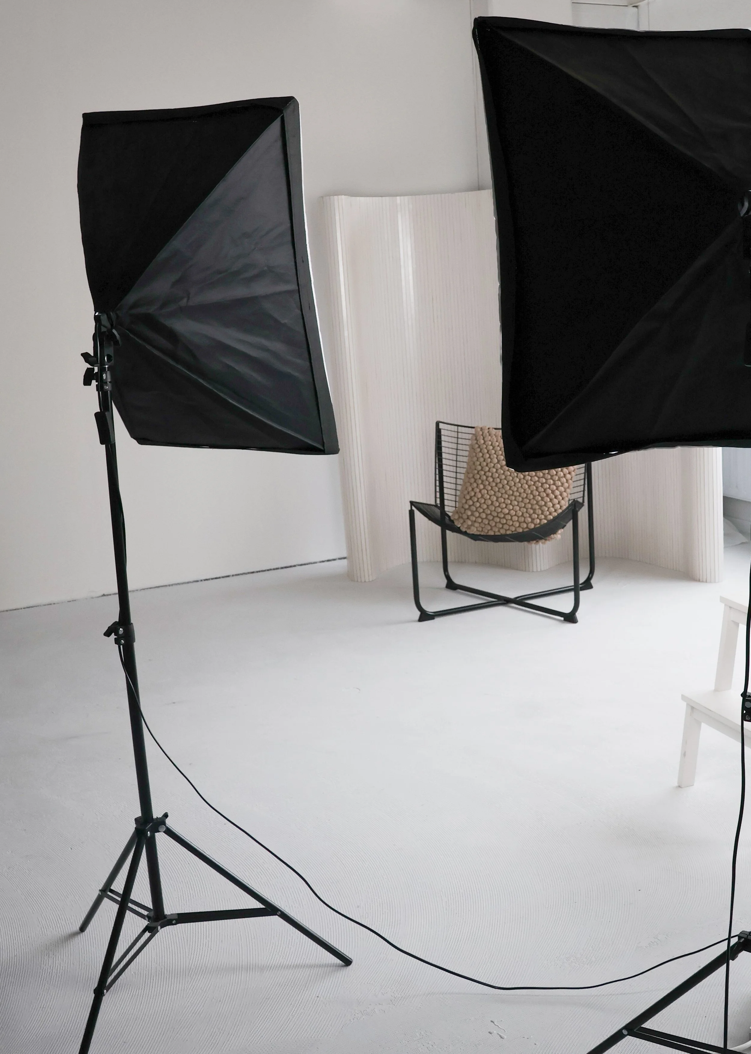 Een fotografeer- of video-opname studio met twee softbox lichten, een zwart metalen stoel met een beige kussen en een witte achtergrond.