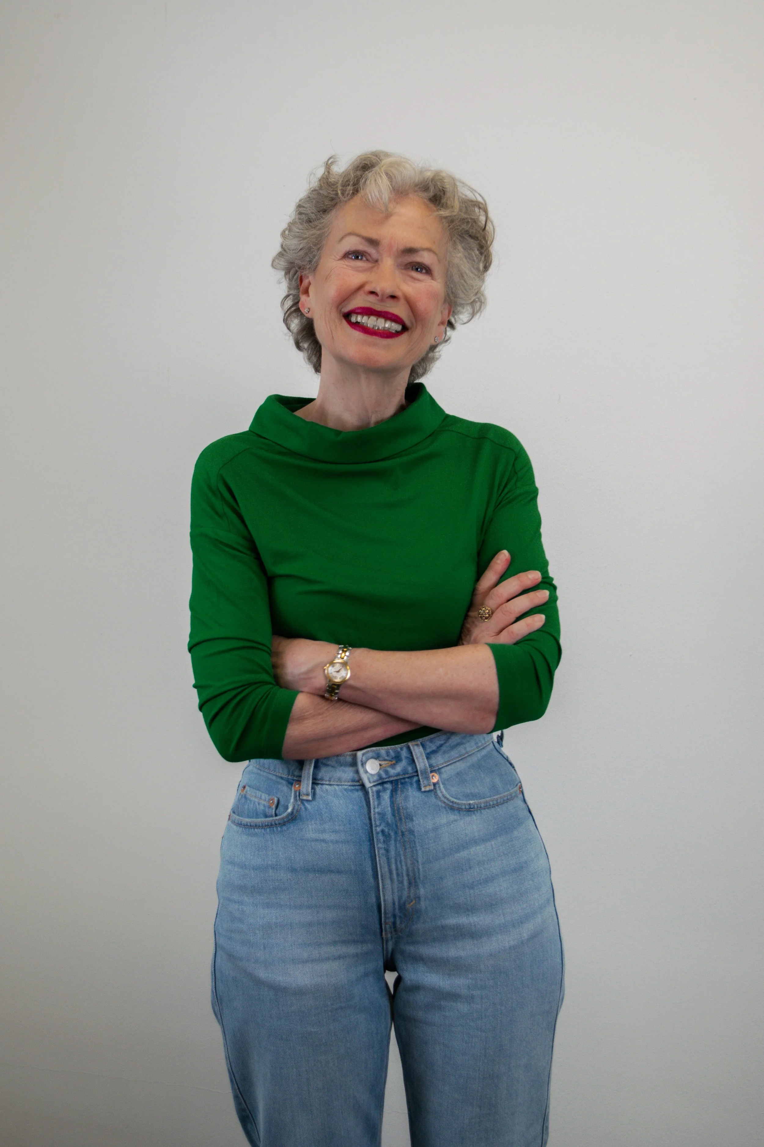 Een vrouw met grijs, krullend haar draagt een groene top en blauwe spijkerbroek, staat lachend met gekruiste armen voor een effen witte achtergrond.