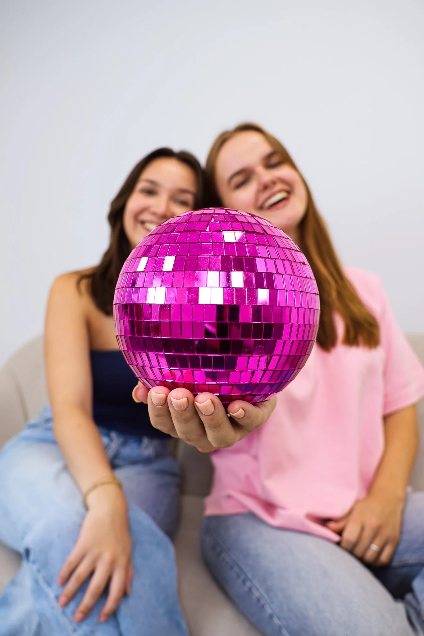 Twee lachende vrouwen zitten op een bank, de vrouw links houdt een roze spiegelbal met kleine spiegelstukjes voor de camera.