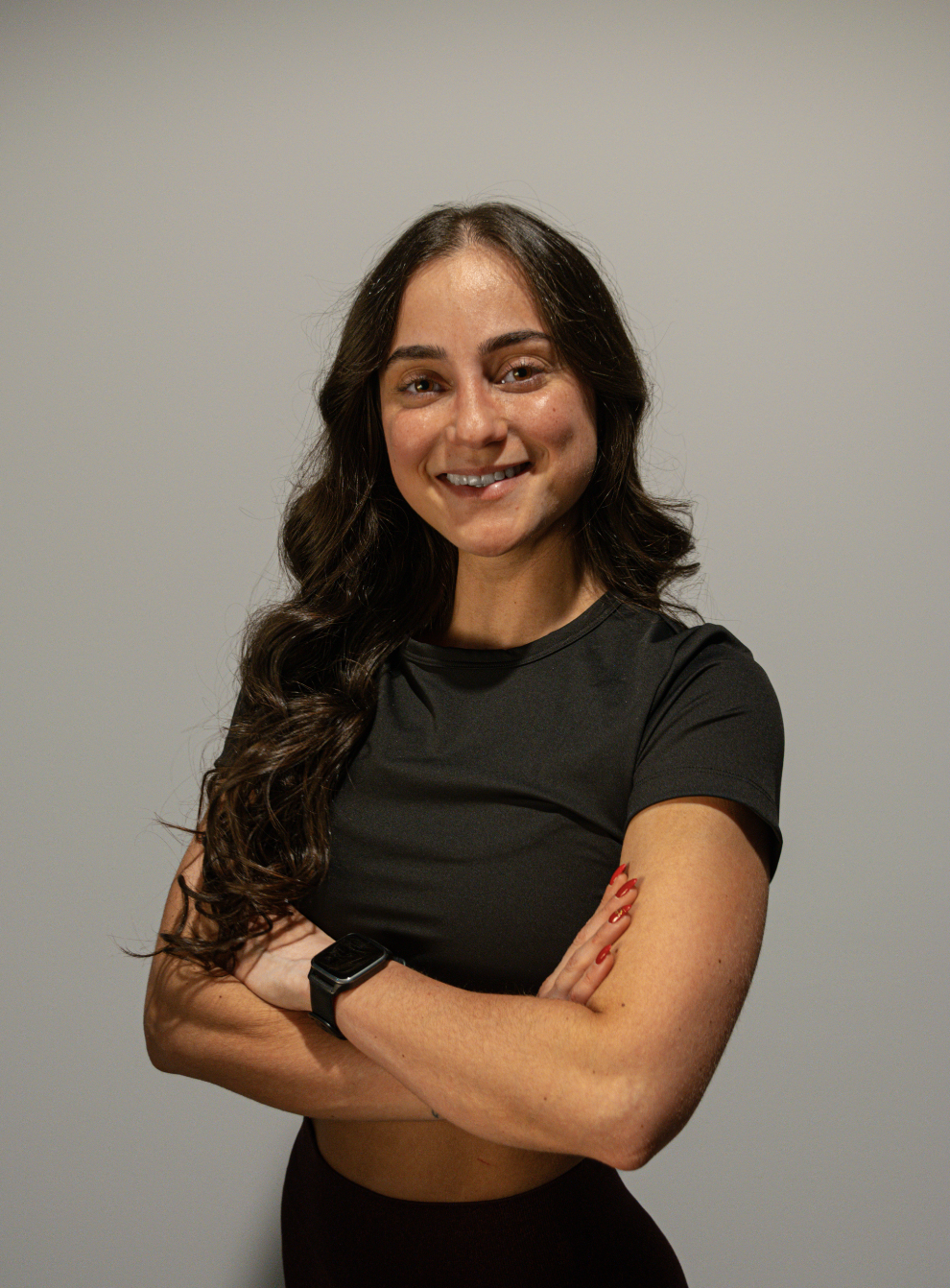Portret van een jonge vrouw met lang, golvend bruin haar, lachend, gekleed in een zwart sportshirt en zwarte sportbroek, met haar armen over elkaar, standing tegen een neutrale grijze achtergrond.