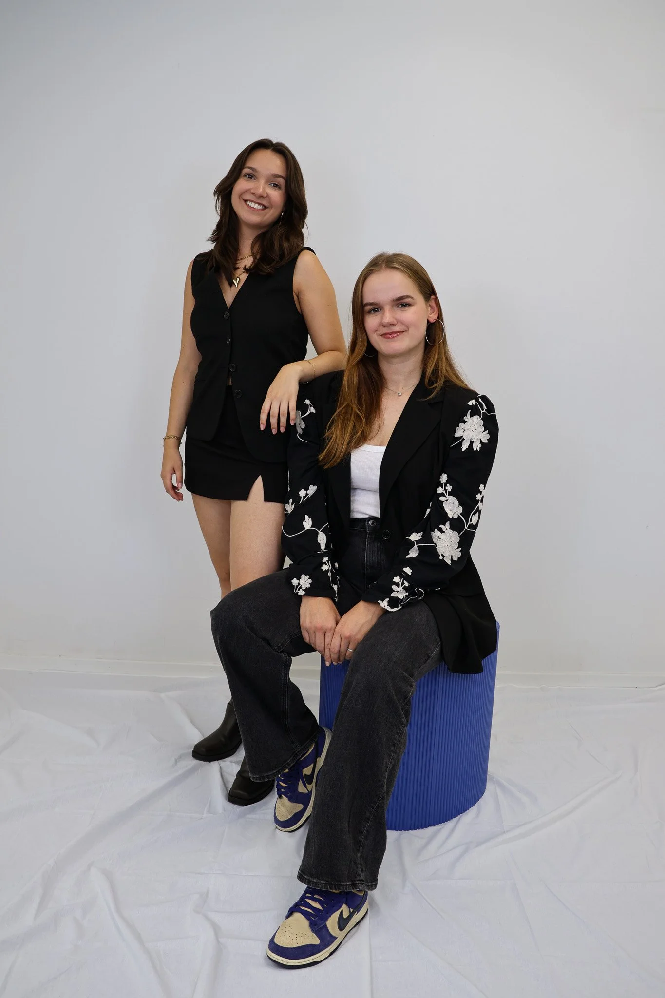 Twee jonge vrouwen in zwarte outfits, een zittend op een blauwe kruk en de andere staand, poseren voor een witte achtergrond.