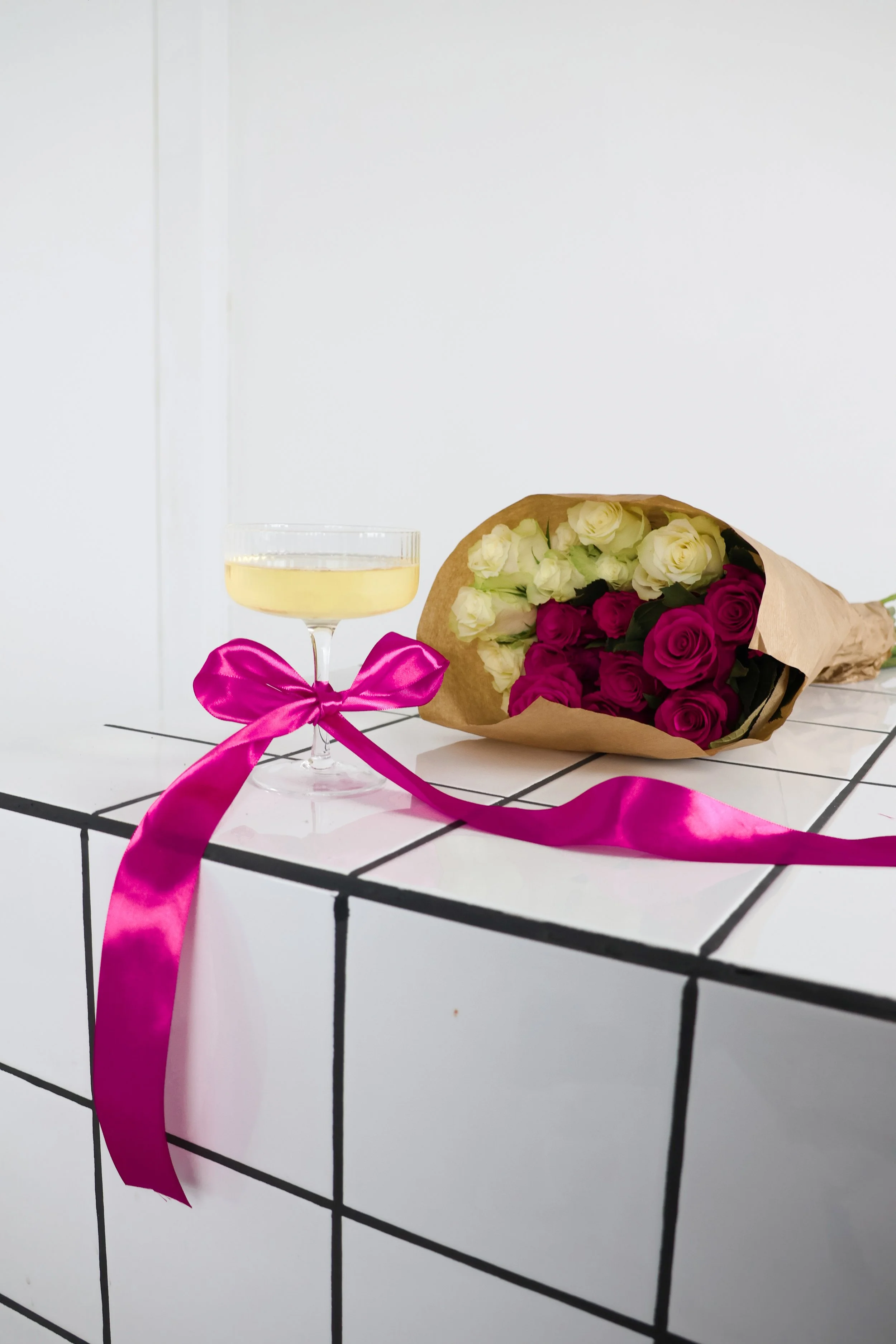 Een witte tafel met zwarte vierkante randen, een glas witte wijn met een roze strik eromheen en een bos bloemen, bestaande uit witte en paarse rozen, ingepakt in bruin papier.