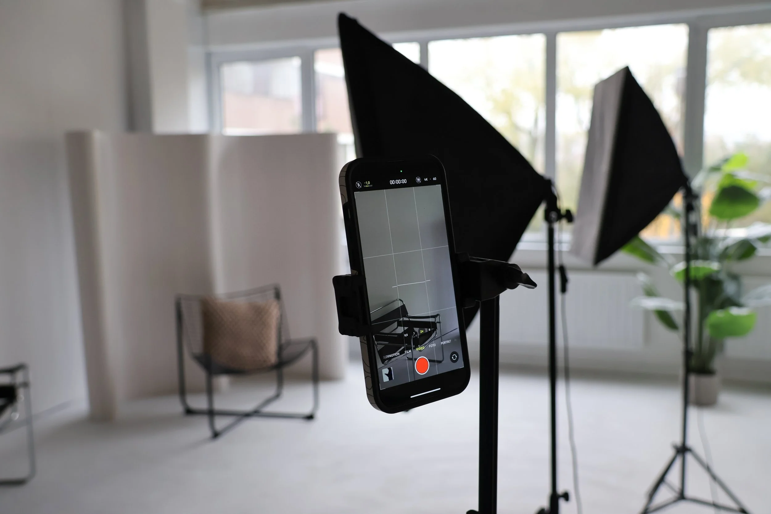 Een smartphone die een video-opname aan het maken is, met een achtergrond van een fotostudio of binnenruimte met grote ramen, een isolatiescherm en planten.