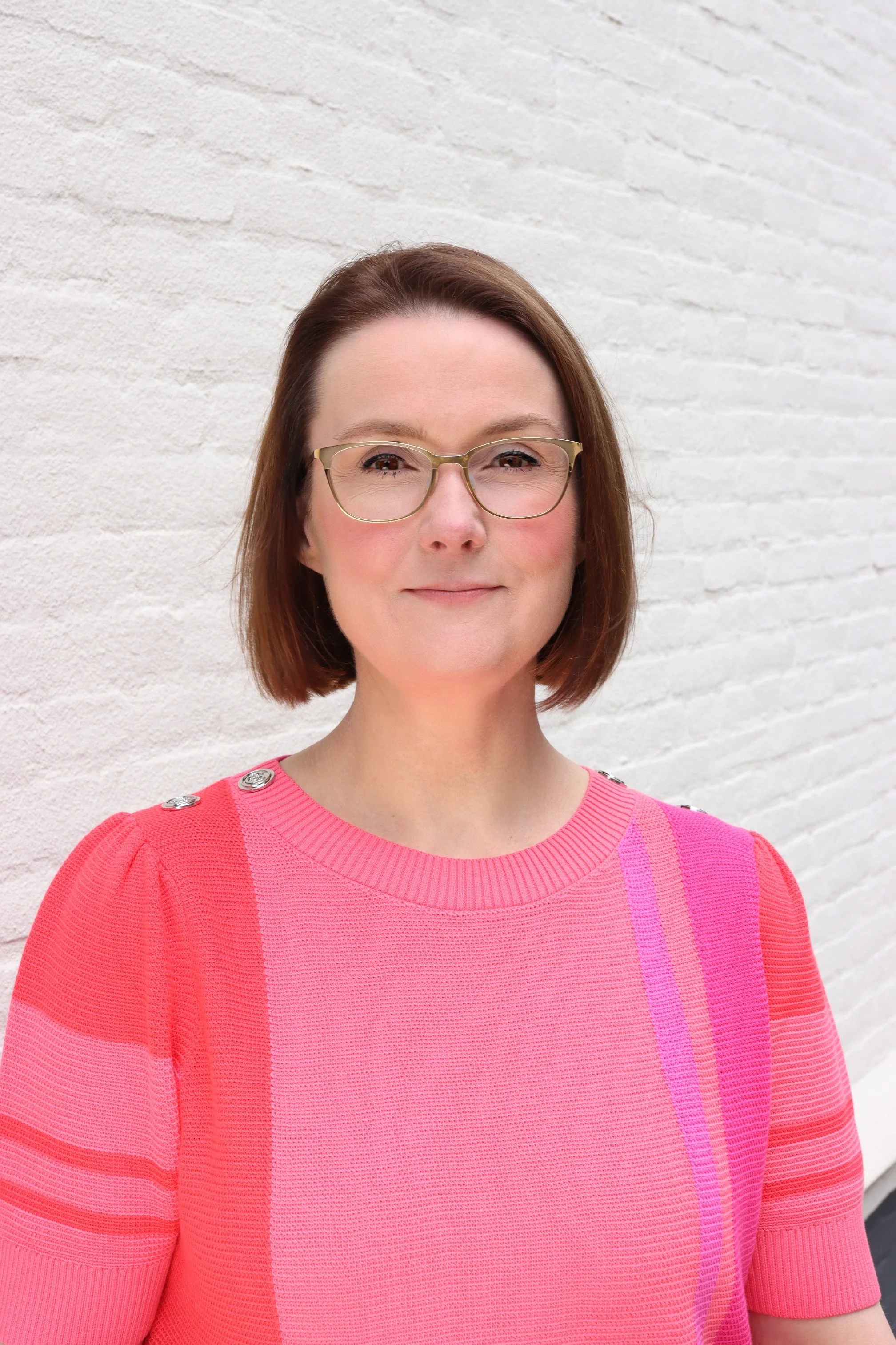 Portret van een vrouw met bruin haar en goudkleurige bril, dragen een roze trui met knopen op de schouder, staand voor een witte bakstenen muur.