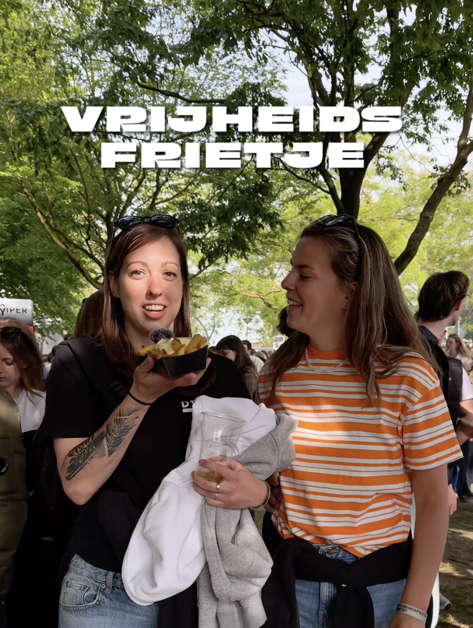 Twee vrouwen praten onder een boom op een festival, een van hen houdt friet en een blauwe bes, de ander lacht.