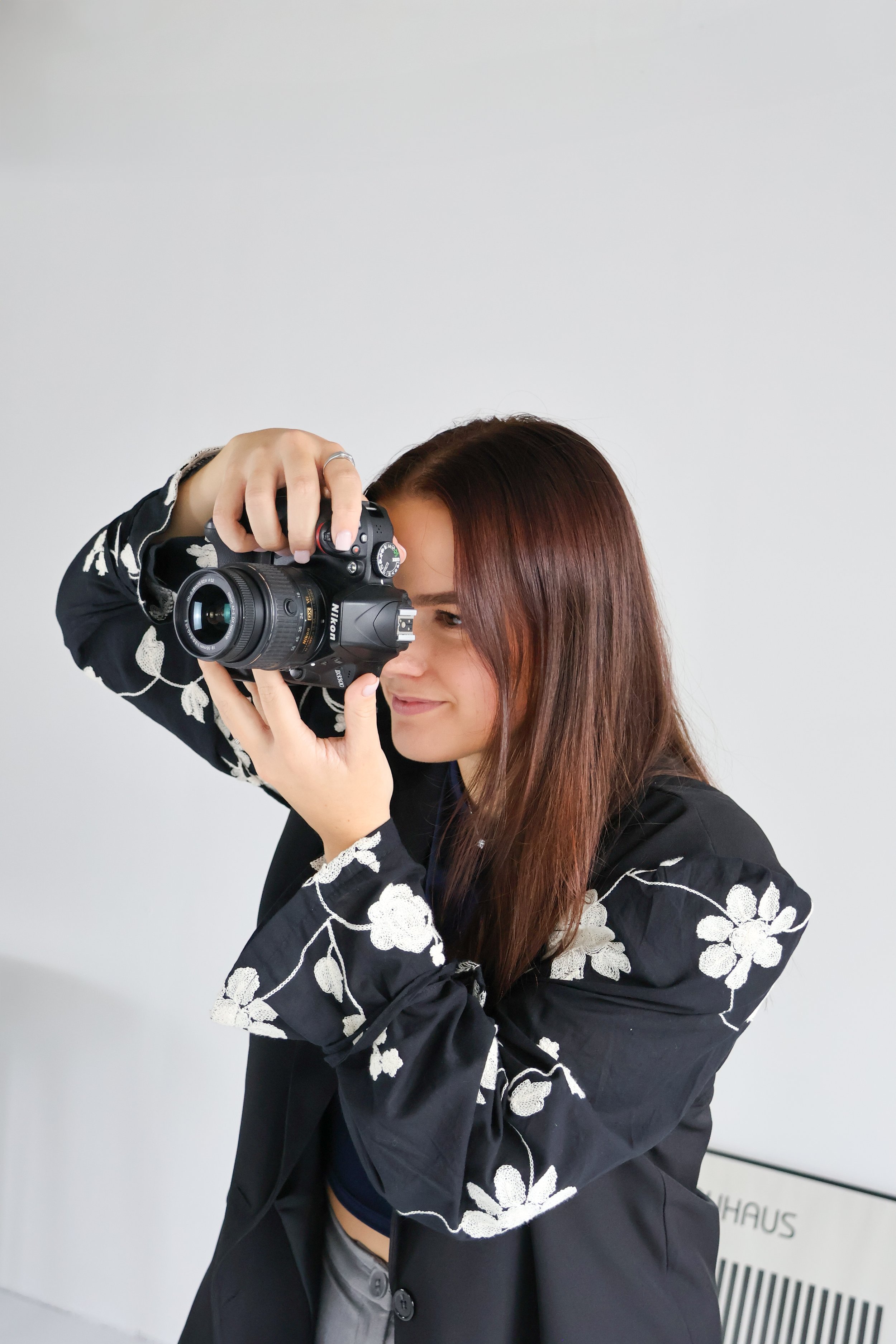Vrouw met bruin haar in een zwarte jas met witte bloemmotieven, die door een camera in een indoor omgeving kijkt.