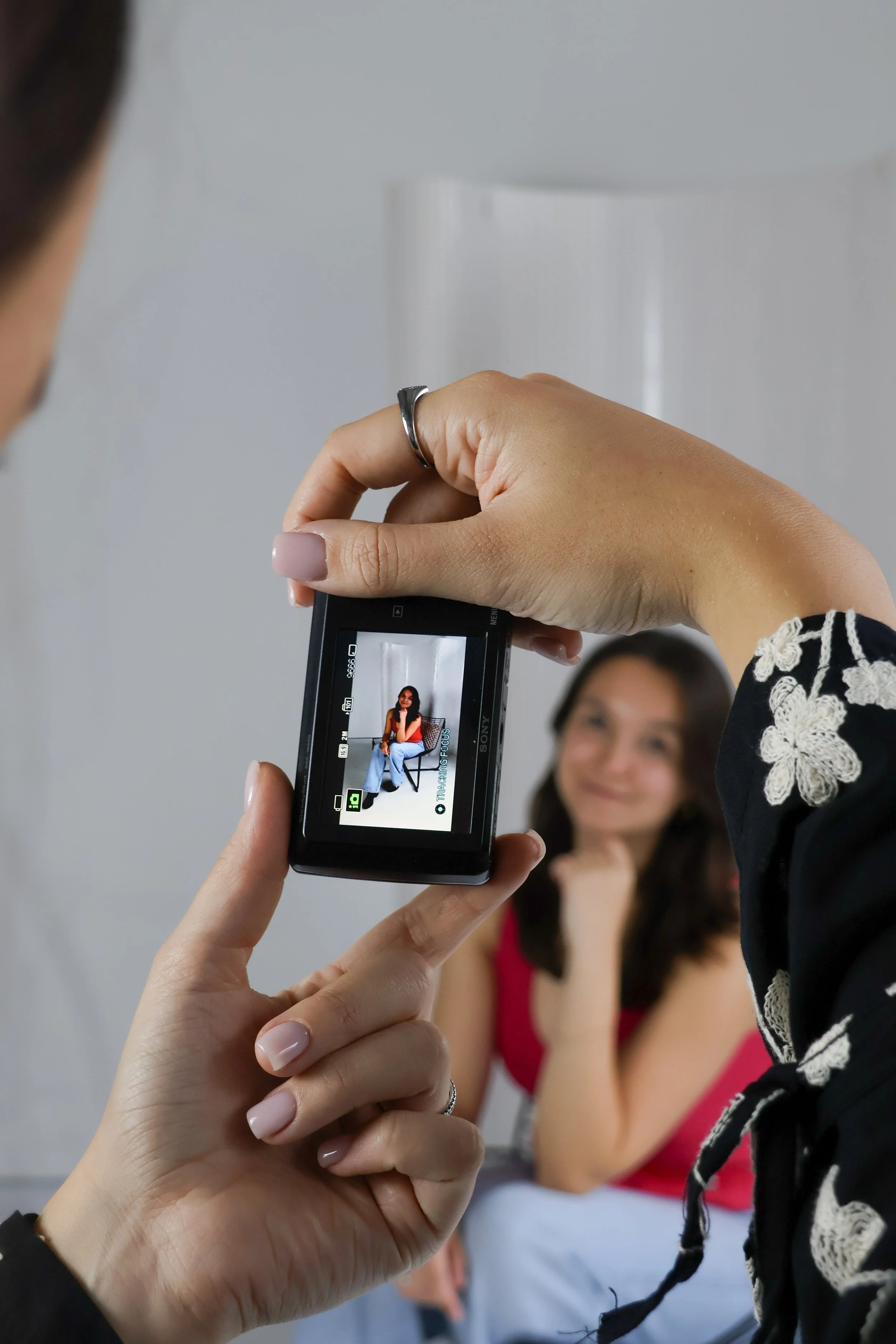 Close-up van een hand die een digitale camera vasthoudt, gericht op een foto van een vrouw die op een stoel zit, terwijl iemand anders een foto neemt. De vrouw op de achtergrond draagt een rode top.