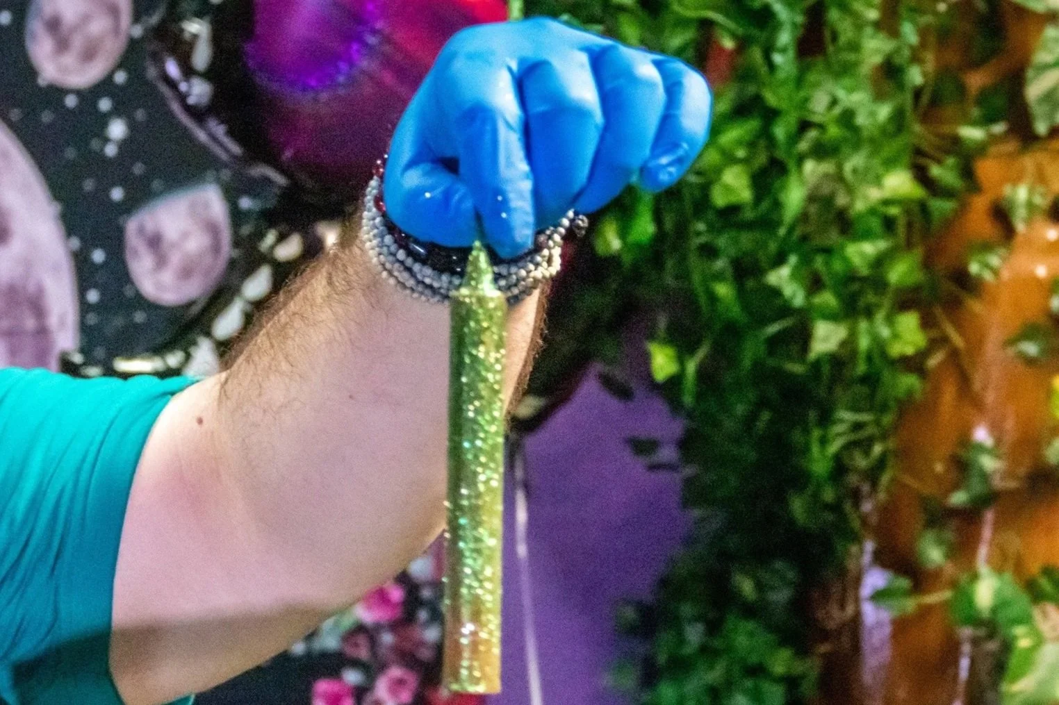 Close-up of a person wearing a blue glove, holding a glittery green wand, with bracelets on their wrist, and a strapless dress with a floral pattern, in front of a background of green foliage and what appears to be a moon or planet illustration.